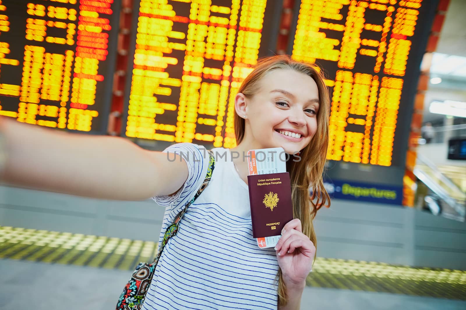 Beautiful young tourist girl in international airport, taking funny selfie with passport and boarding pass near flight information board