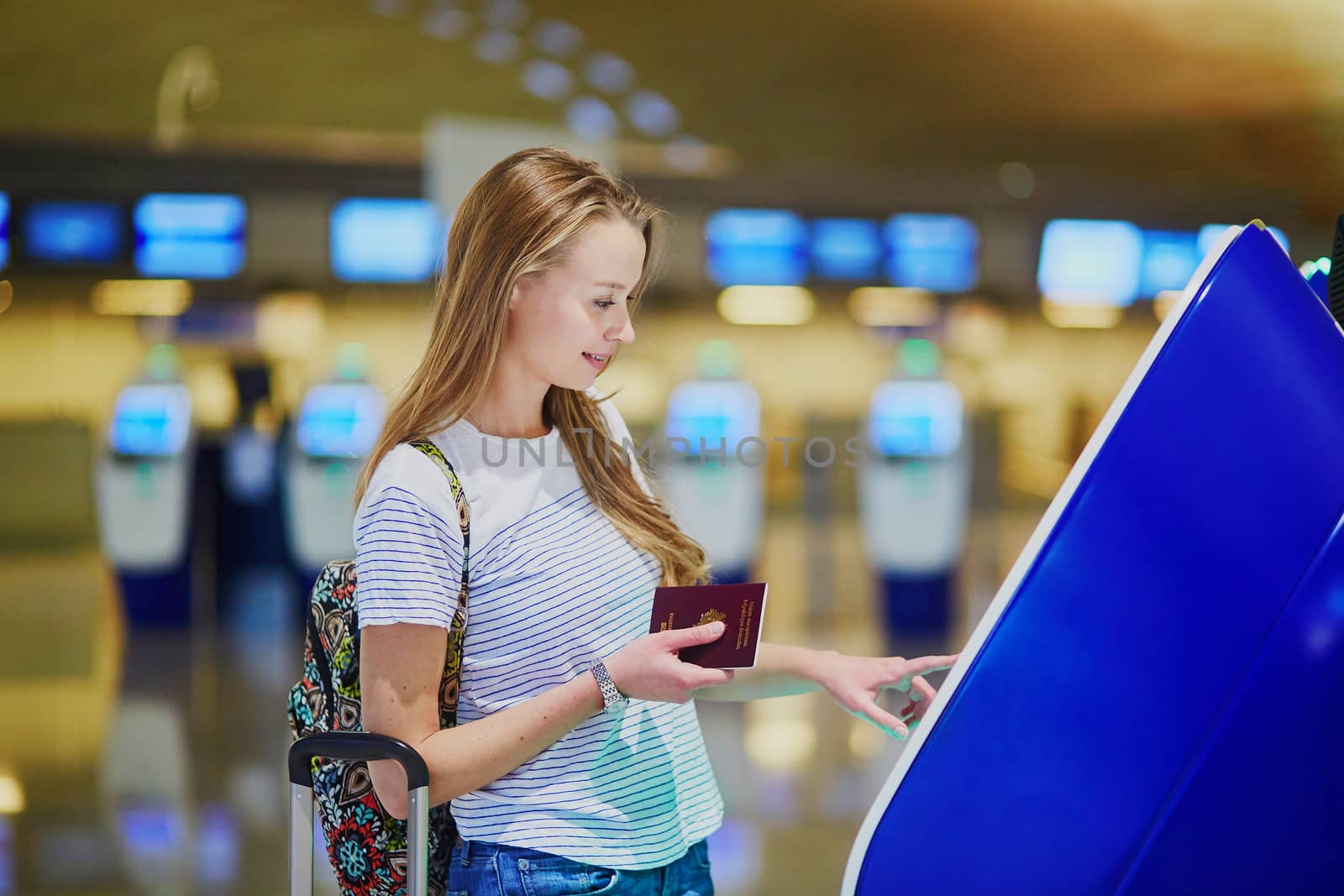 Beautiful young tourist girl with backpack and carry on luggage in international airport, doing self check-in