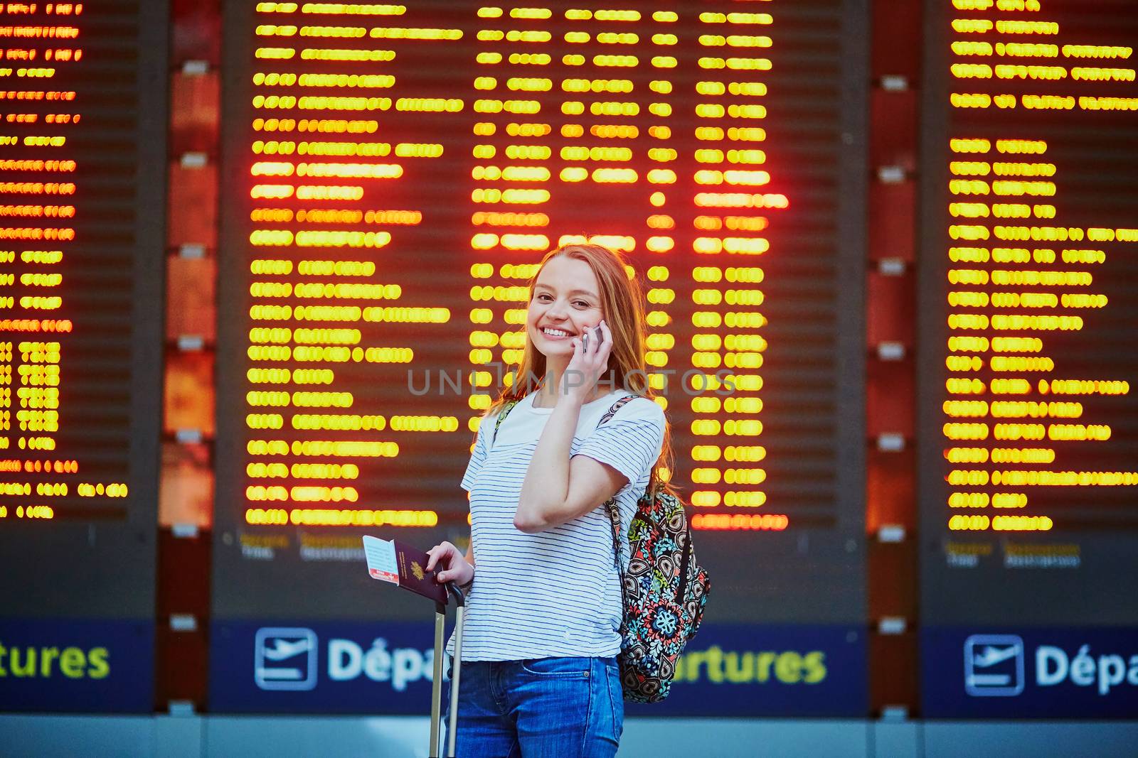 Beautiful young tourist girl with backpack and carry on luggage in international airport, near flight information board