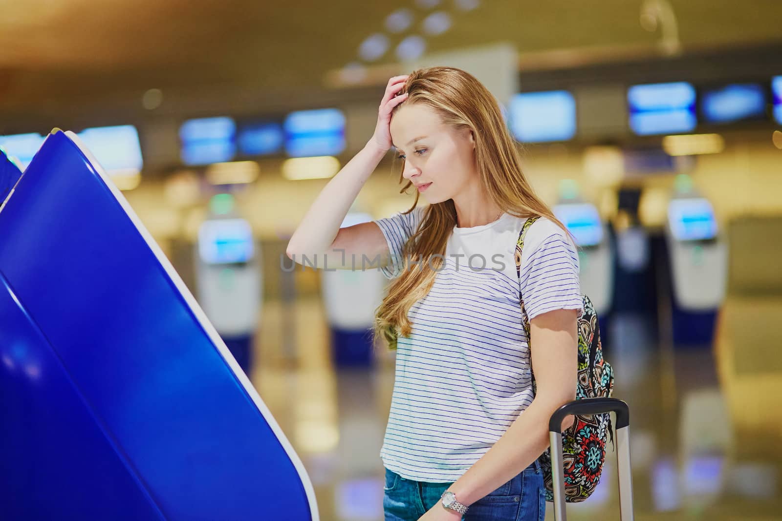 Young tourist girl with backpack and carry on luggage in international airport, doing self check-in, looking upset and worried. Delayed or missed flight concept