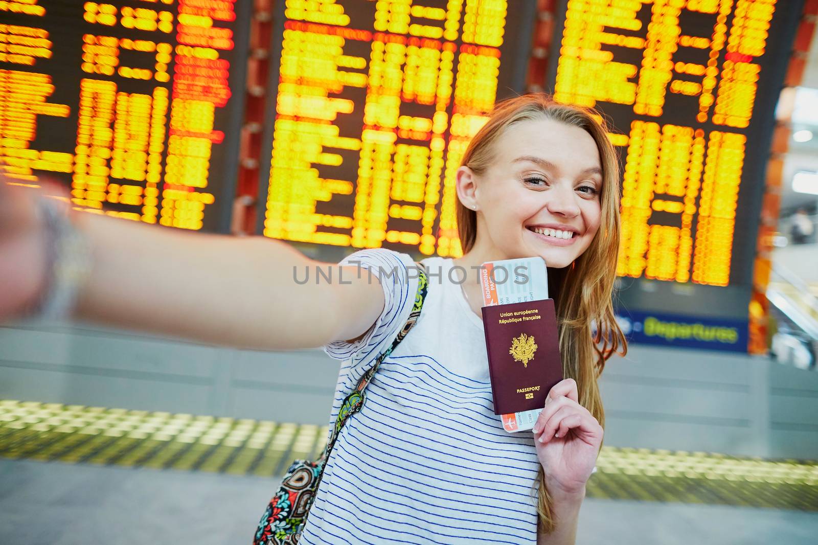 Beautiful young tourist girl in international airport, taking funny selfie with passport and boarding pass near flight information board