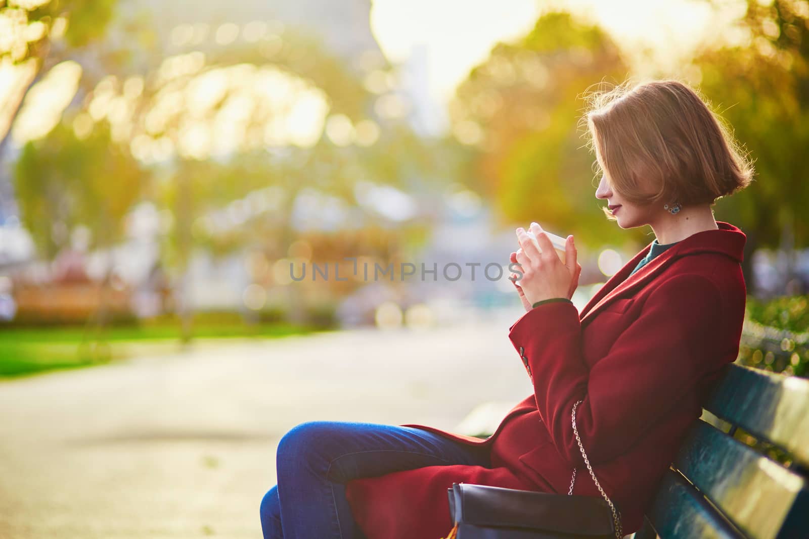 Beautiful young French woman sitting on the bench and drinking coffee near the Eiffel tower in Paris on a fall day