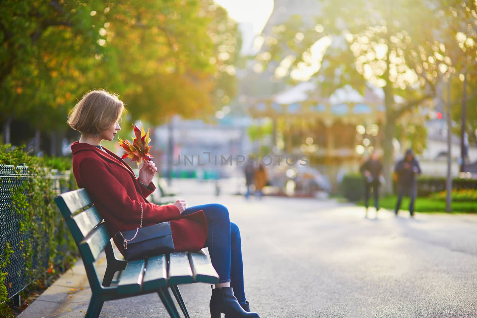 Beautiful young French woman with bunch of colorful autumn leaves sitting on the bench near the Eiffel tower in Paris on a fall day