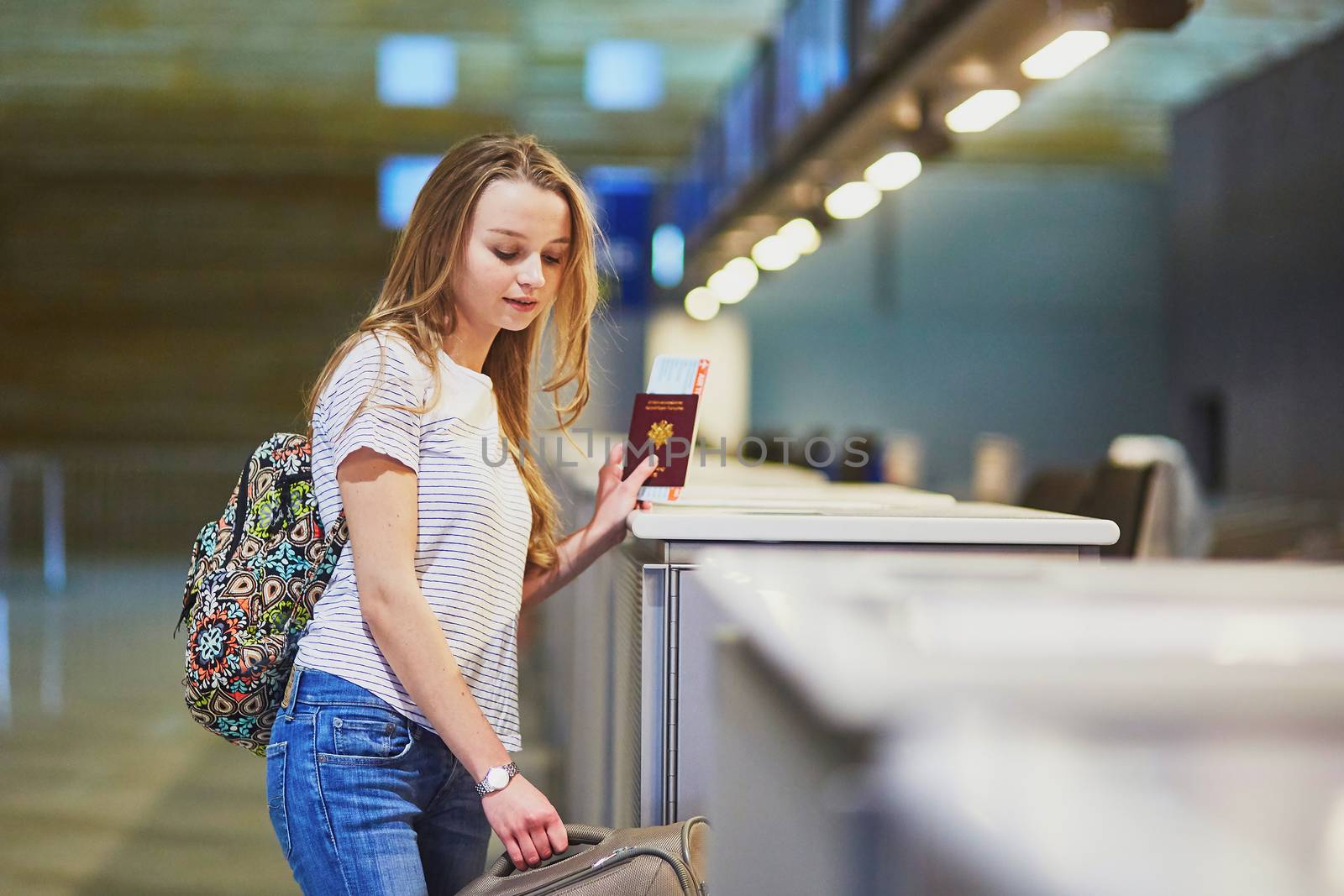 Beautiful young tourist girl with backpack and carry on luggage in international airport at check-in counter, giving her passport to an officer and waiting for her boarding pass