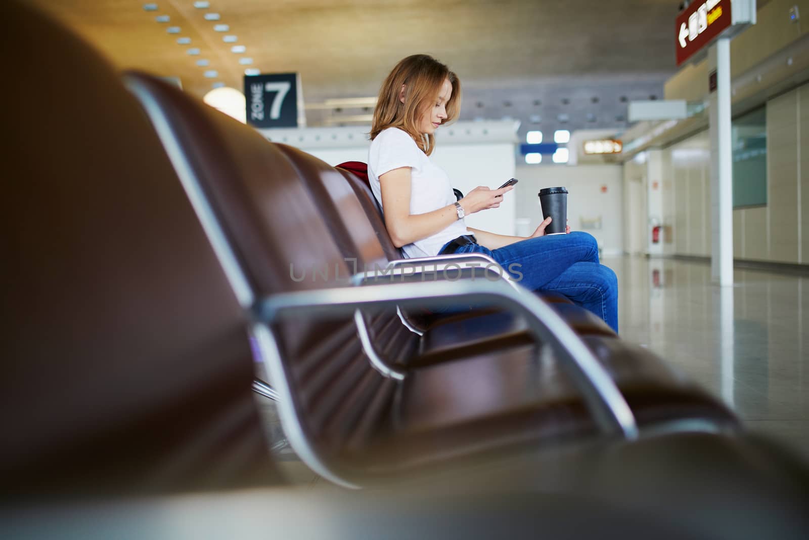 Young woman in international airport with luggage and coffee to go, waiting for her flight