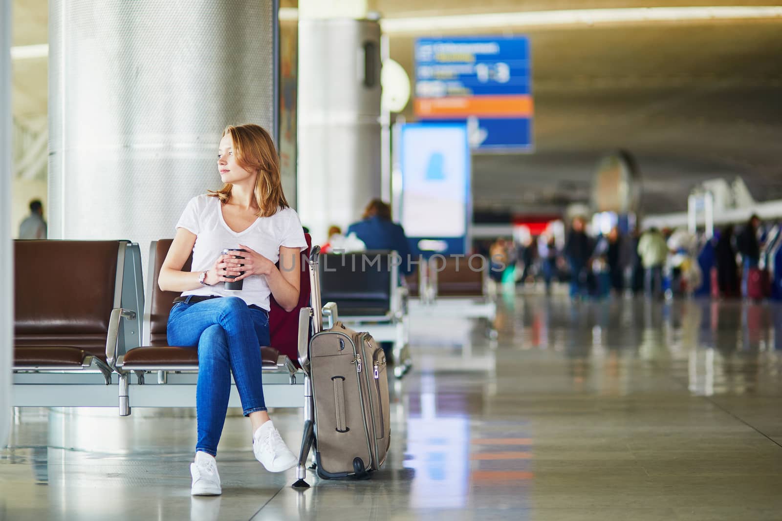 Young woman in international airport with luggage and coffee to go, waiting for her flight