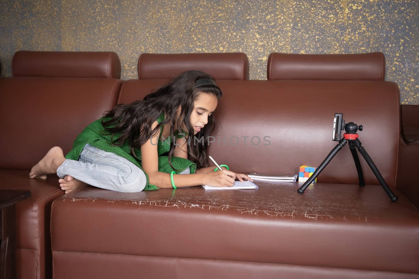 Concept of improper, lazy and way of studying during homeschooling or e-learning and make kids sleepy or boredom, young girl busy in writing by looking into mobile on sofa during covid-19 pandemic.