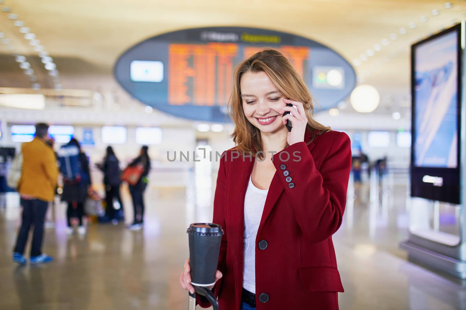 Young woman in international airport with luggage and coffee to go, speaking on the phone and waiting for her flight