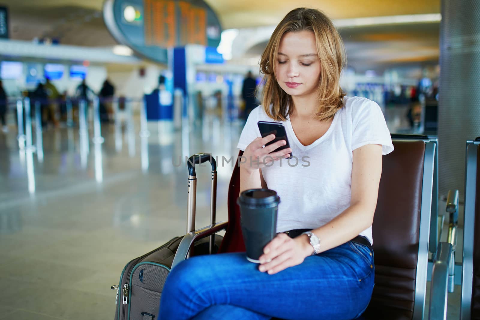 Young woman in international airport with luggage and coffee to go, waiting for her flight and looking at her phone