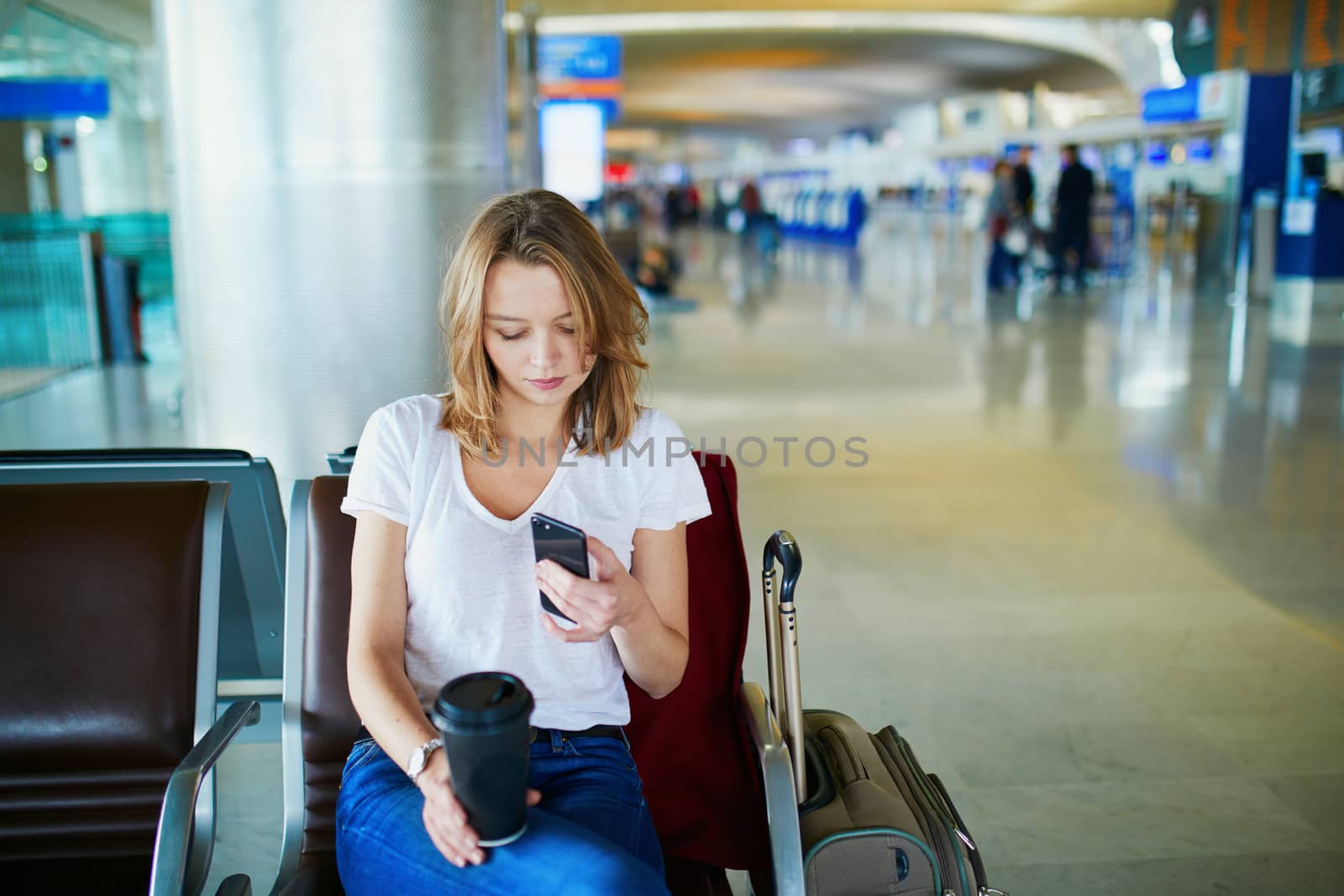 Young woman in international airport with luggage and coffee to go, waiting for her flight and looking at her phone