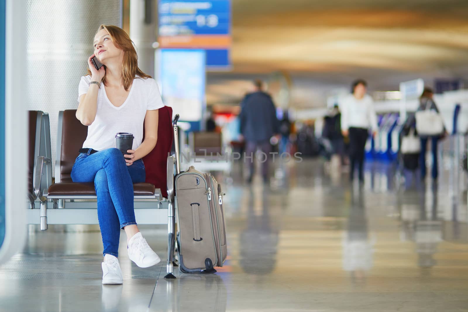 Young woman in international airport with luggage and coffee to go, waiting for her flight and speaking on the phone