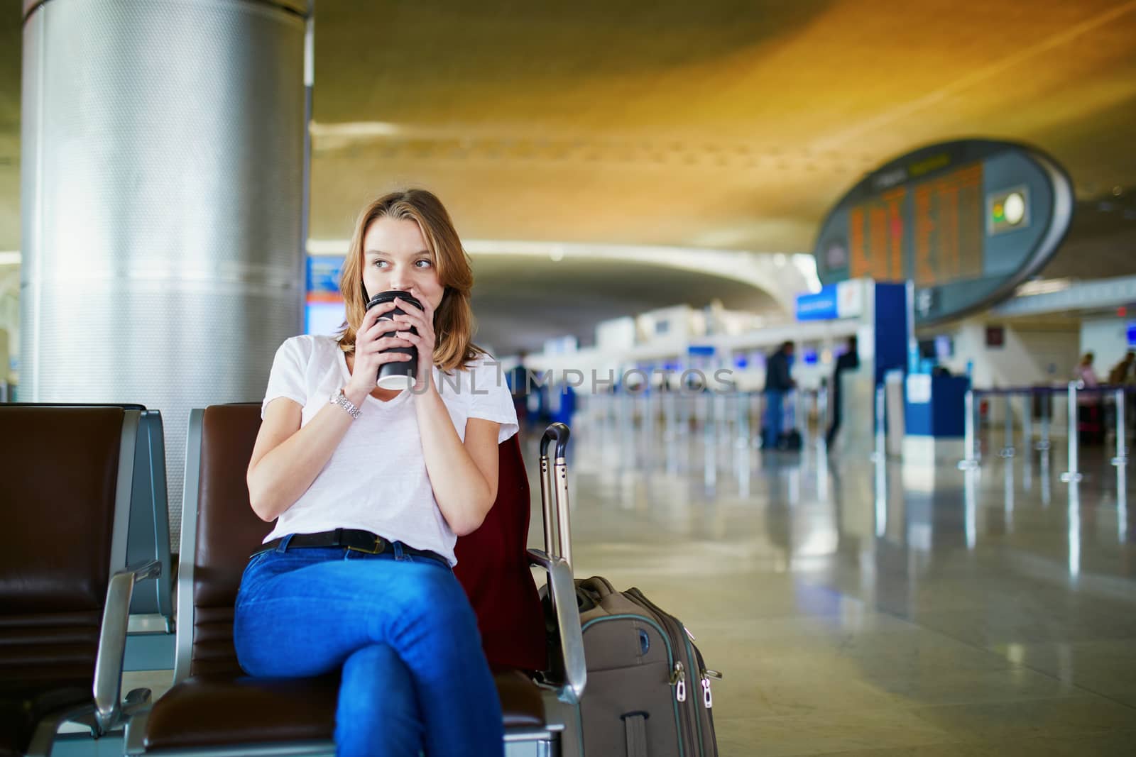 Young woman in international airport with luggage and coffee to go, waiting for her flight