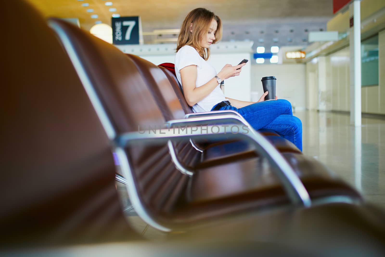 Young woman in international airport with luggage and coffee to go, waiting for her flight and looking at her phone