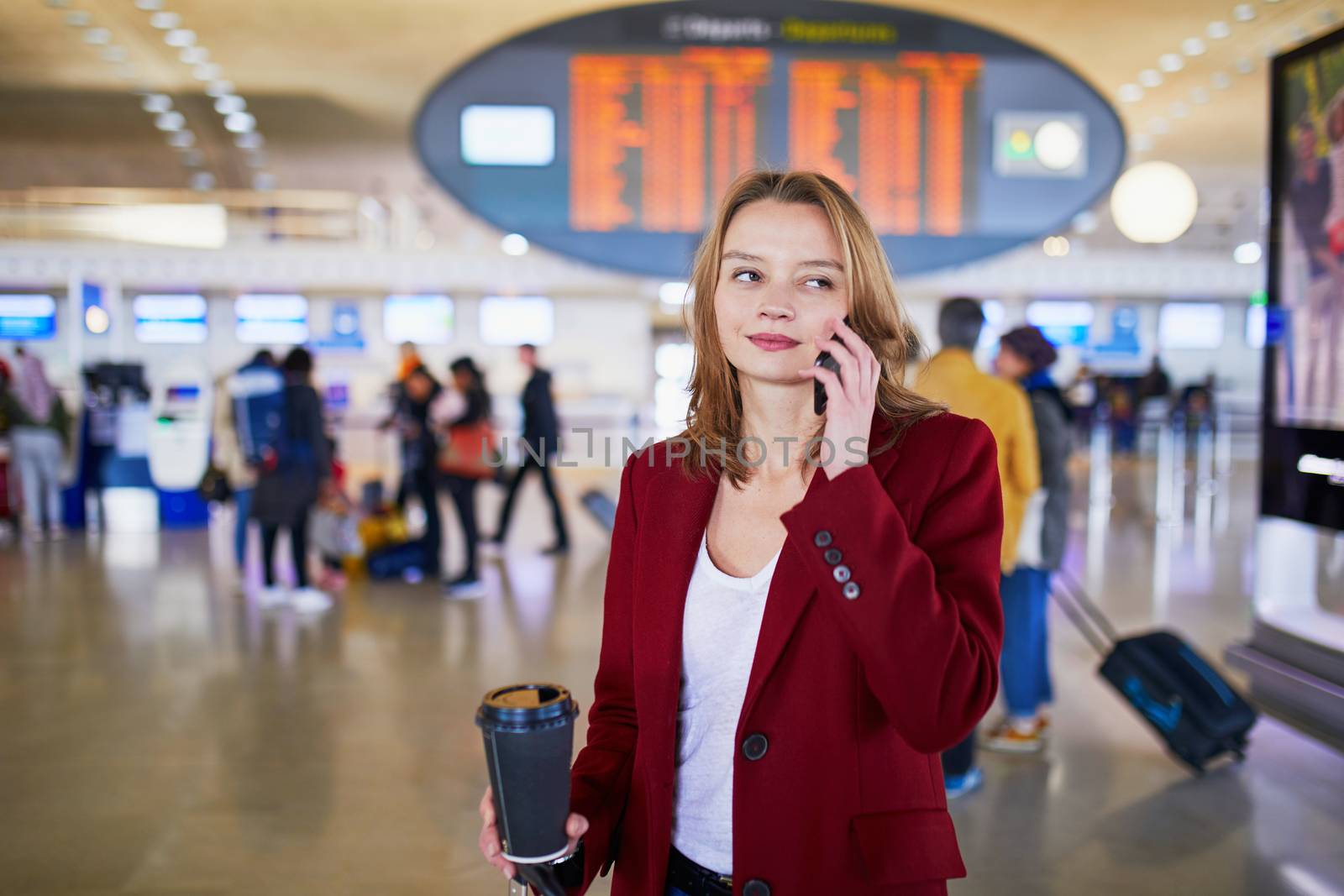 Young woman in international airport with luggage and coffee to go, speaking on the phone and waiting for her flight