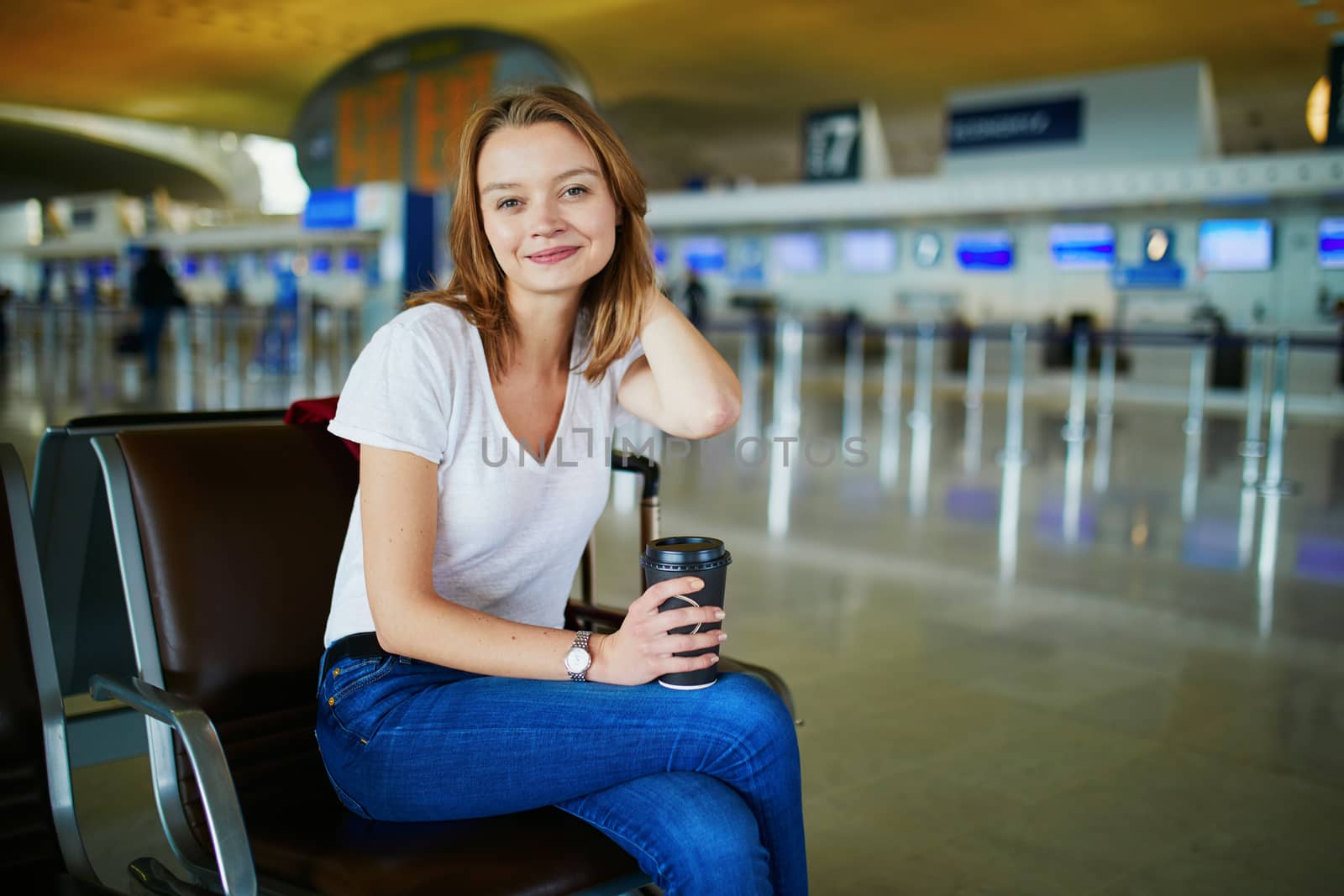 Young woman in international airport with luggage and coffee to go, waiting for her flight