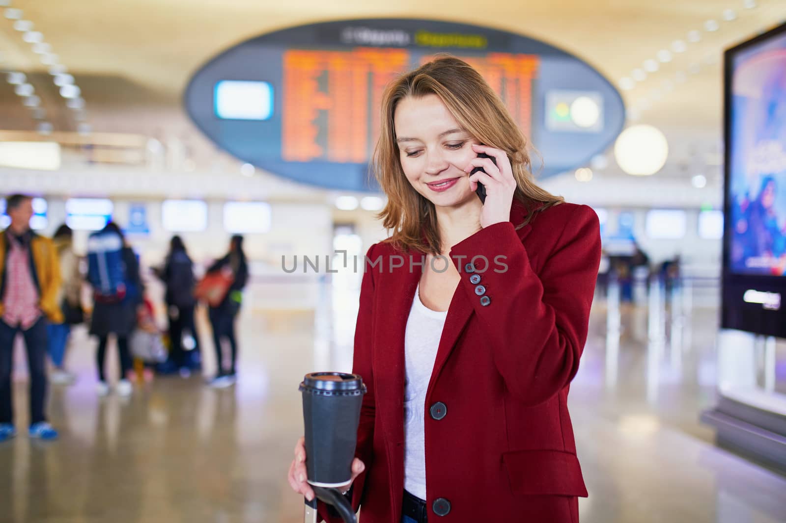 Young woman in international airport with luggage and coffee to go, speaking on the phone and waiting for her flight