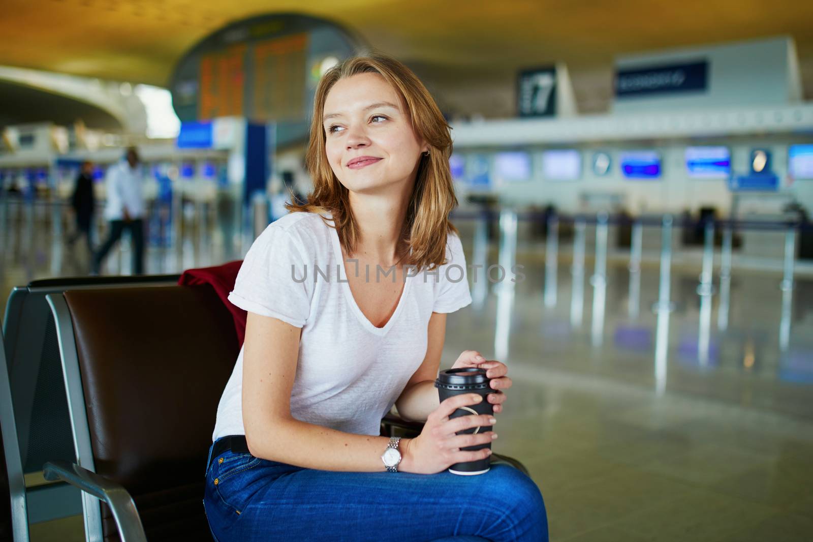 Young woman in international airport with luggage and coffee to go, waiting for her flight