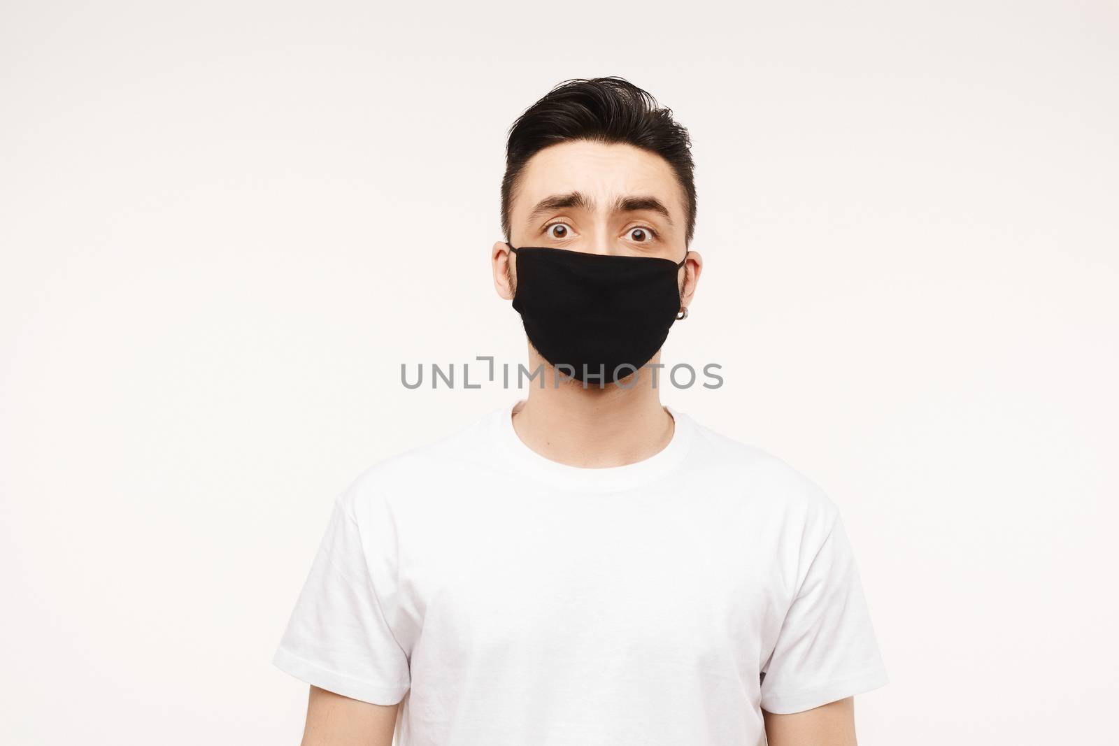 Scared man in a medical mask looks in camera. Scared young man in fear of coronavirus wearing white t-shirt and protective medical mask against viruses and infections, isolated at white background with copy space.