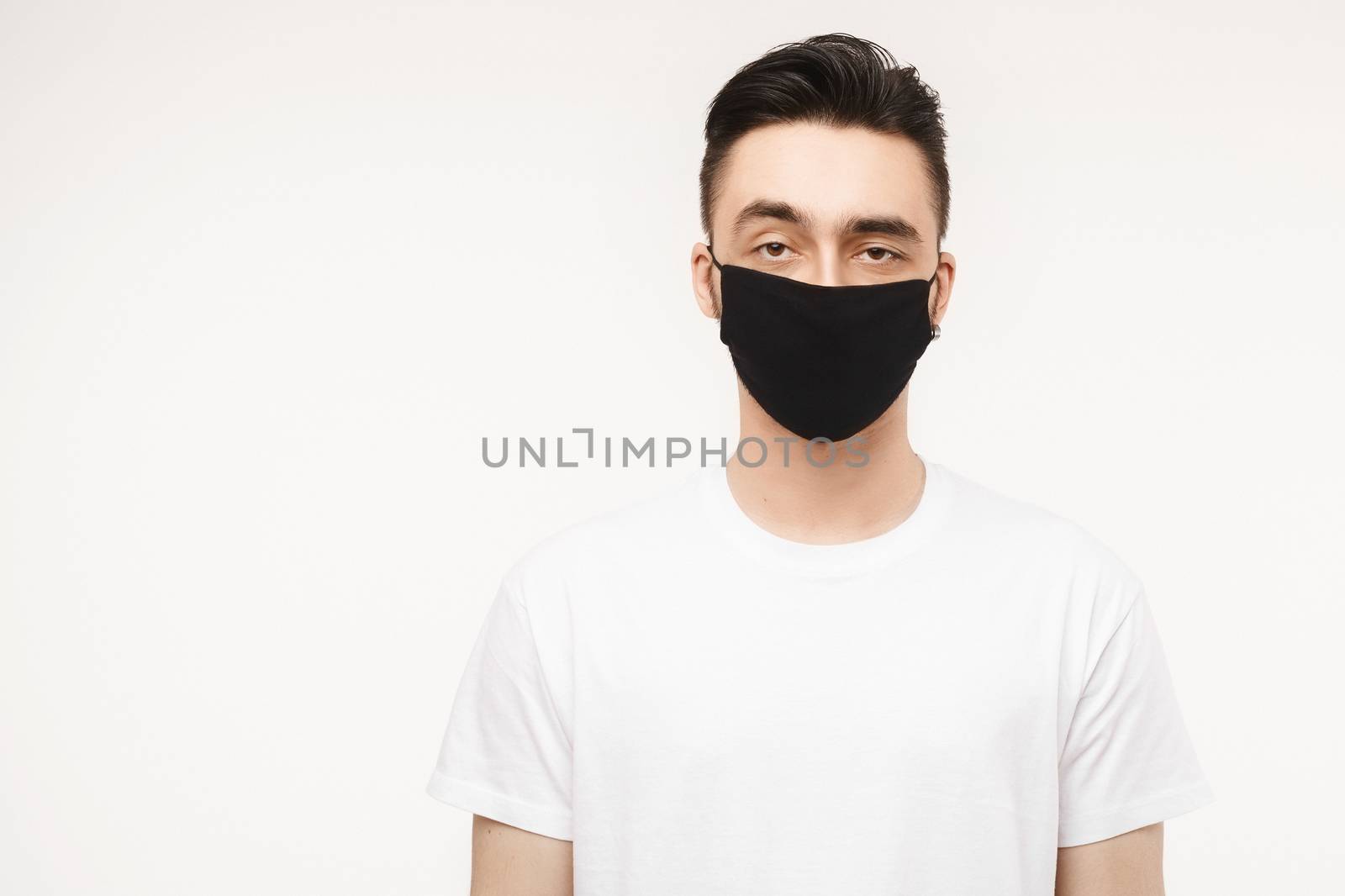 Young man in white t-shirt and black protective mask isolated on white background in studio. Healthy lifestyle concept, seasonal illness, and seasonal flu concept. Man wearing medical mask to prevent infection, airborne respiratory illness such as flu, 2019-nCoV.