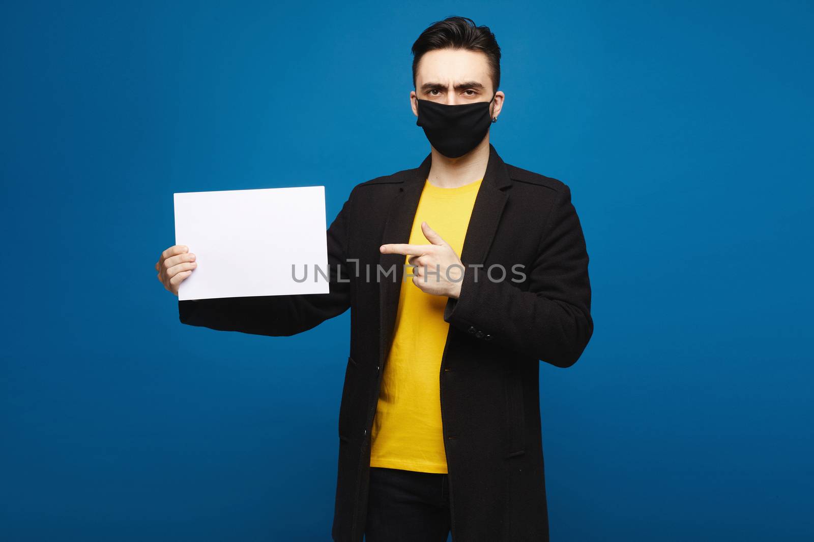Young handsome man holding blank sheet of paper, isolated at the blue background. Young guy pointing at a sheet of the paper sheet and looking at the camera. Promotion concept.
