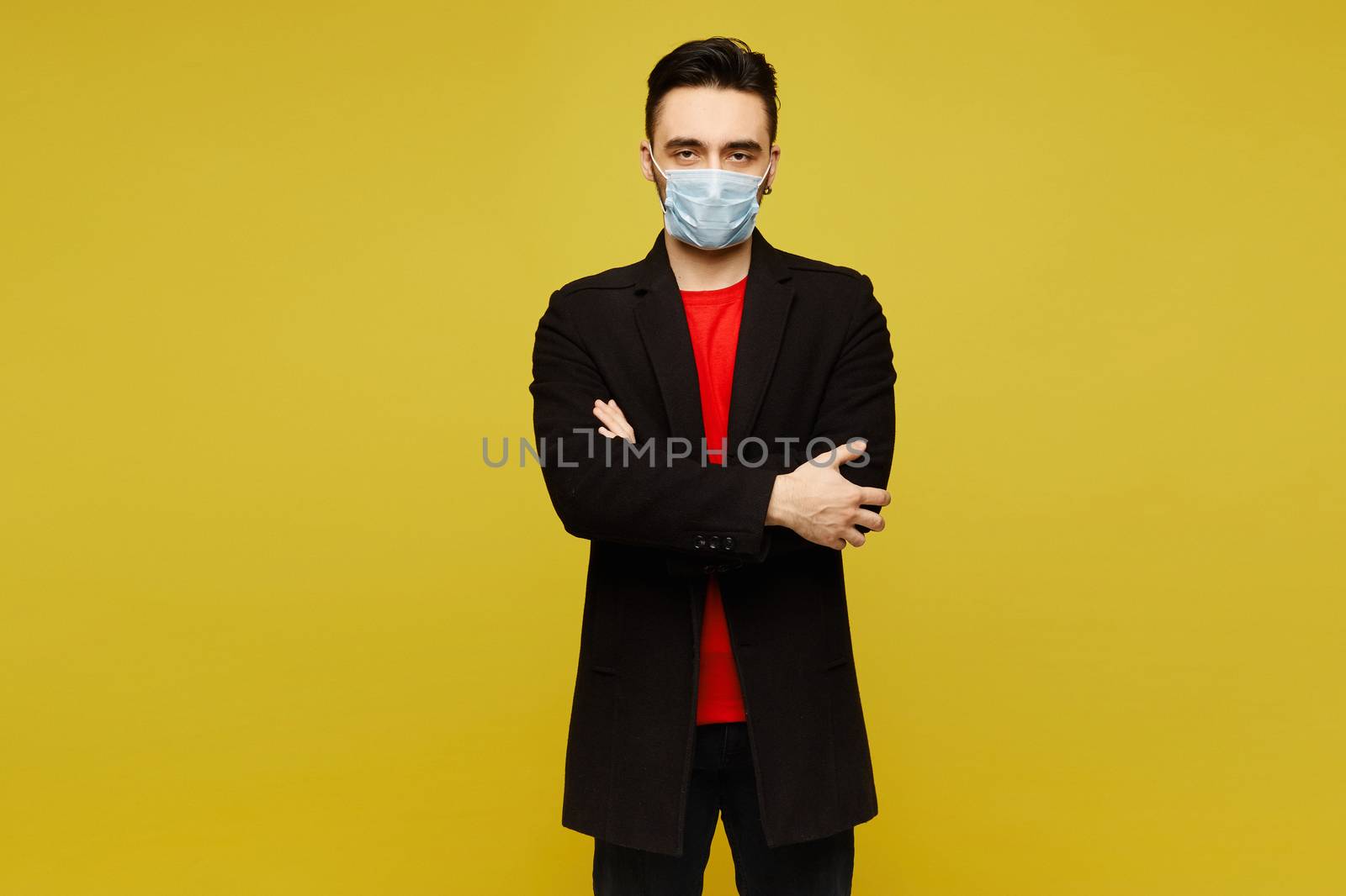 Young man in a red t-shirt and coat wears a protective medical mask and posing with folded hands, isolated in a yellow background. Advertisement mockup.