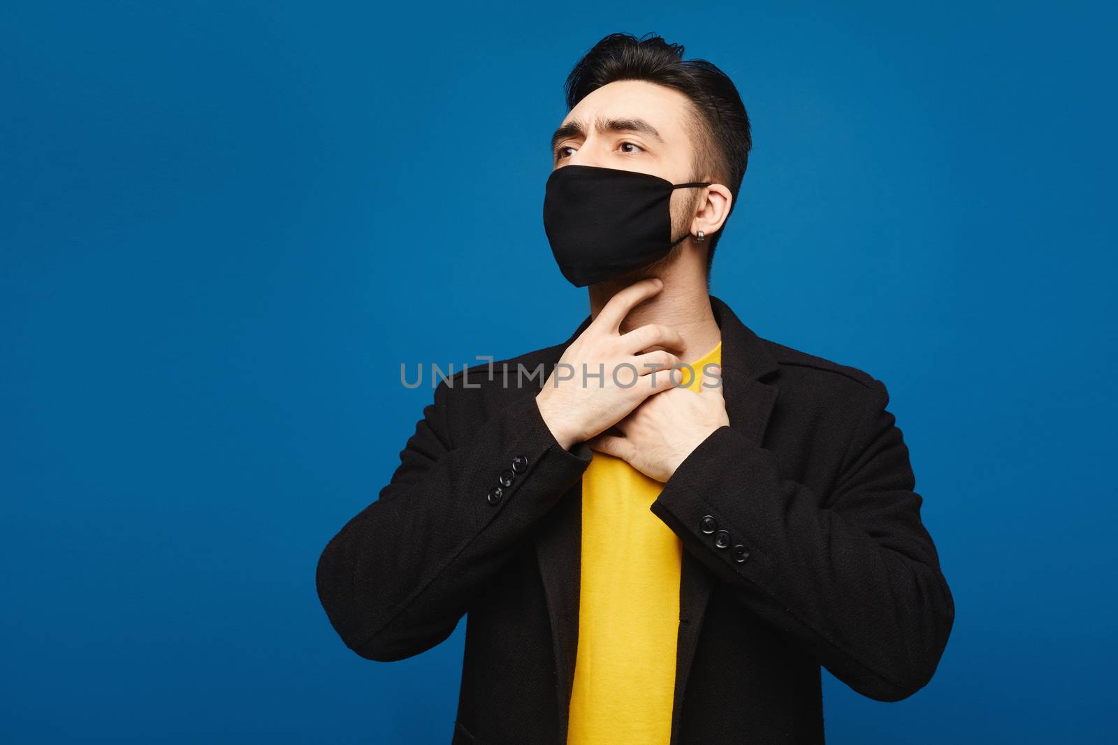 Young man in the black protective mask with throat sore. Handsome men holds hands on his neck because of throat pain. Inflamed throat. Portrait of sick man in medical mask touching painful neck, medical concept.