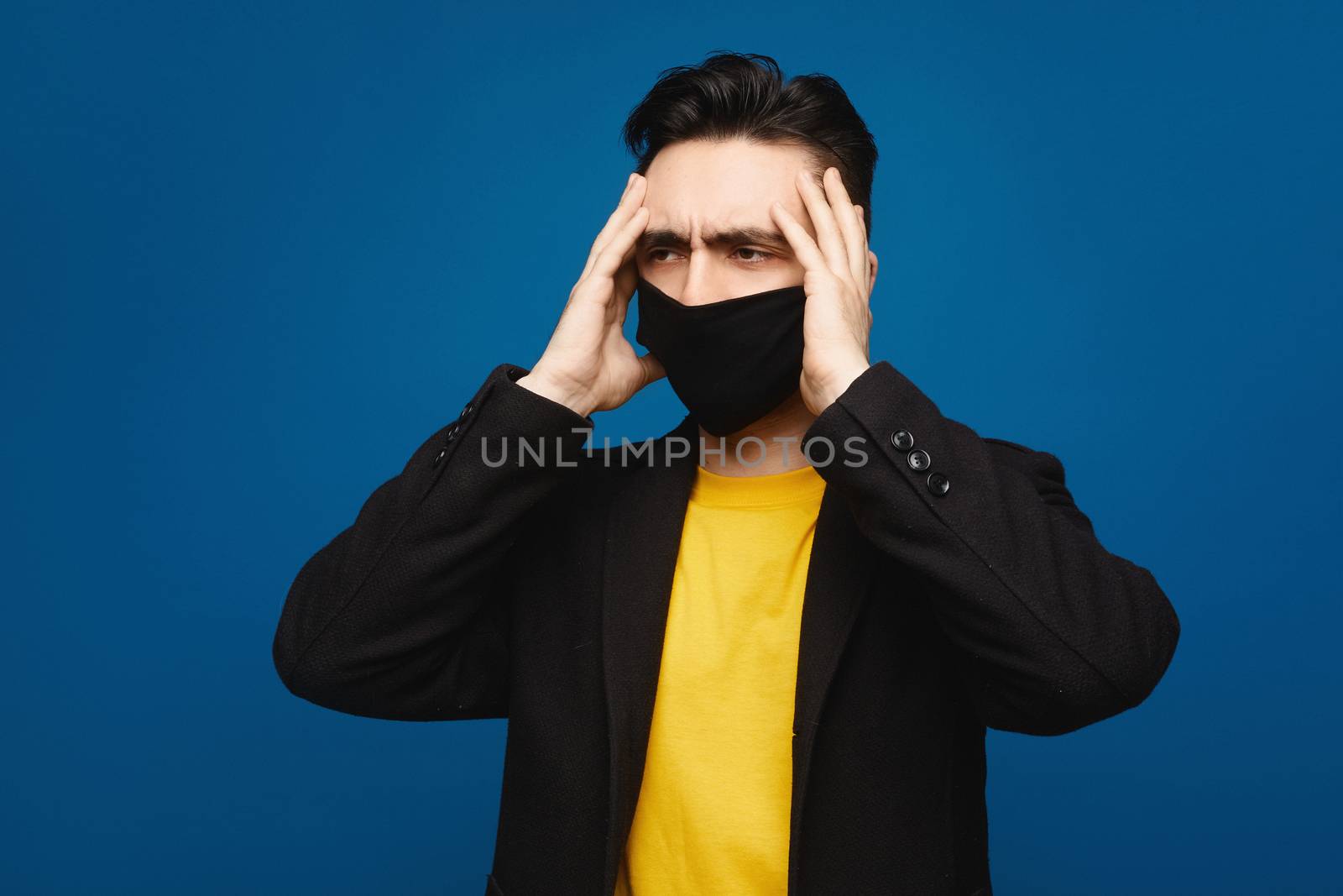 Portrait of young ill man in medical protective mask on face suffering from headache and weakness, isolated at blue background. Typical symptoms of coronavirus COVID-19 or flu. Healthcare concept.
