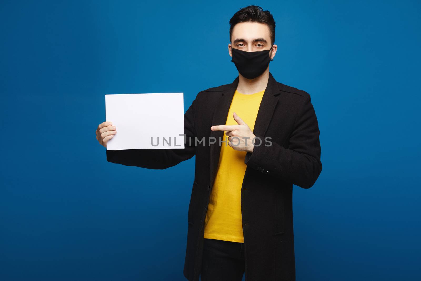 Young handsome man in black protective mask holding blank sheet of paper and pointing on it, isolated at the blue background. Promotion concept. Healthcare concept.
