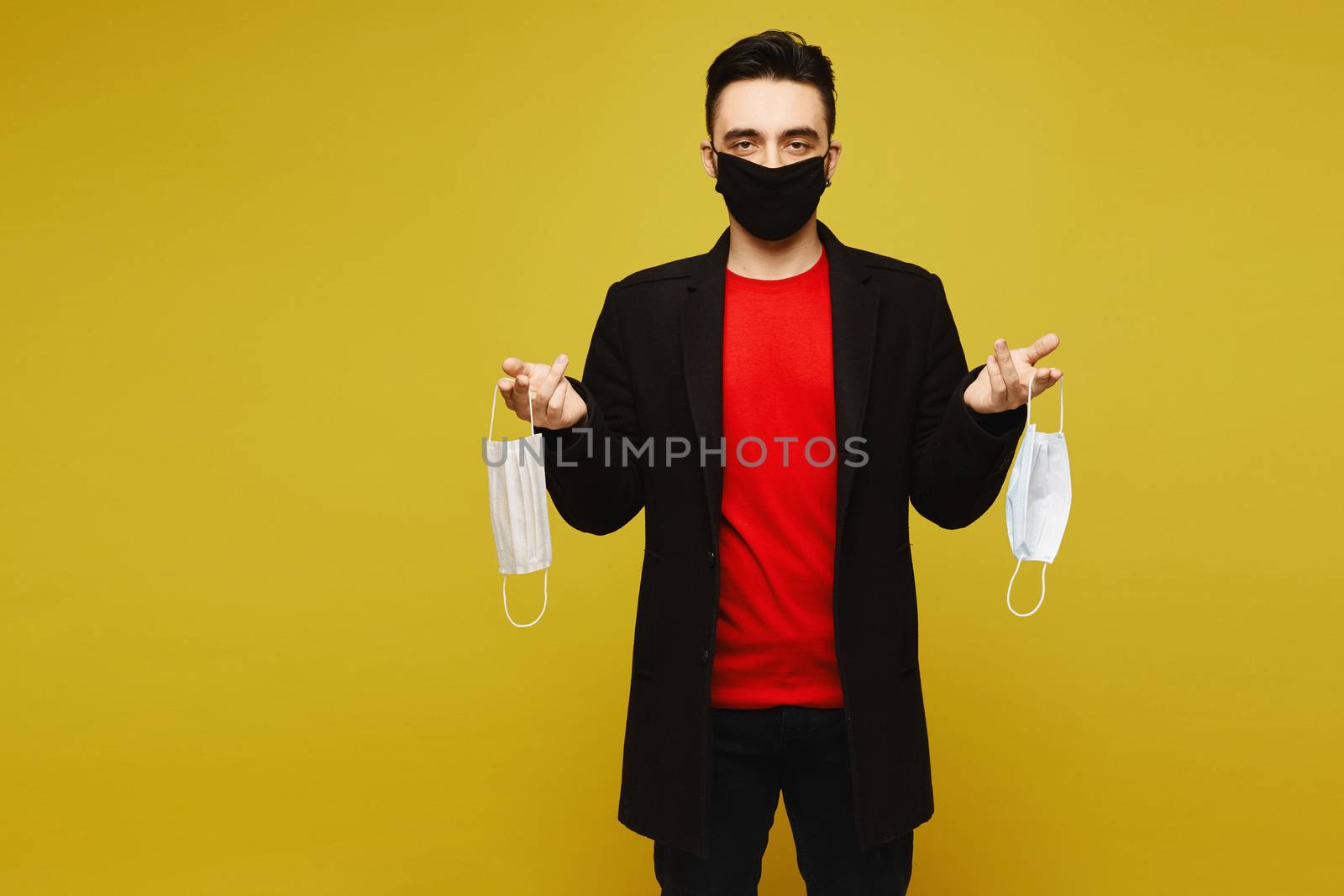 Young man in a protective medical mask holding a pair of protective masks in hands, isolated in a yellow background. Medical advertisement mockup.