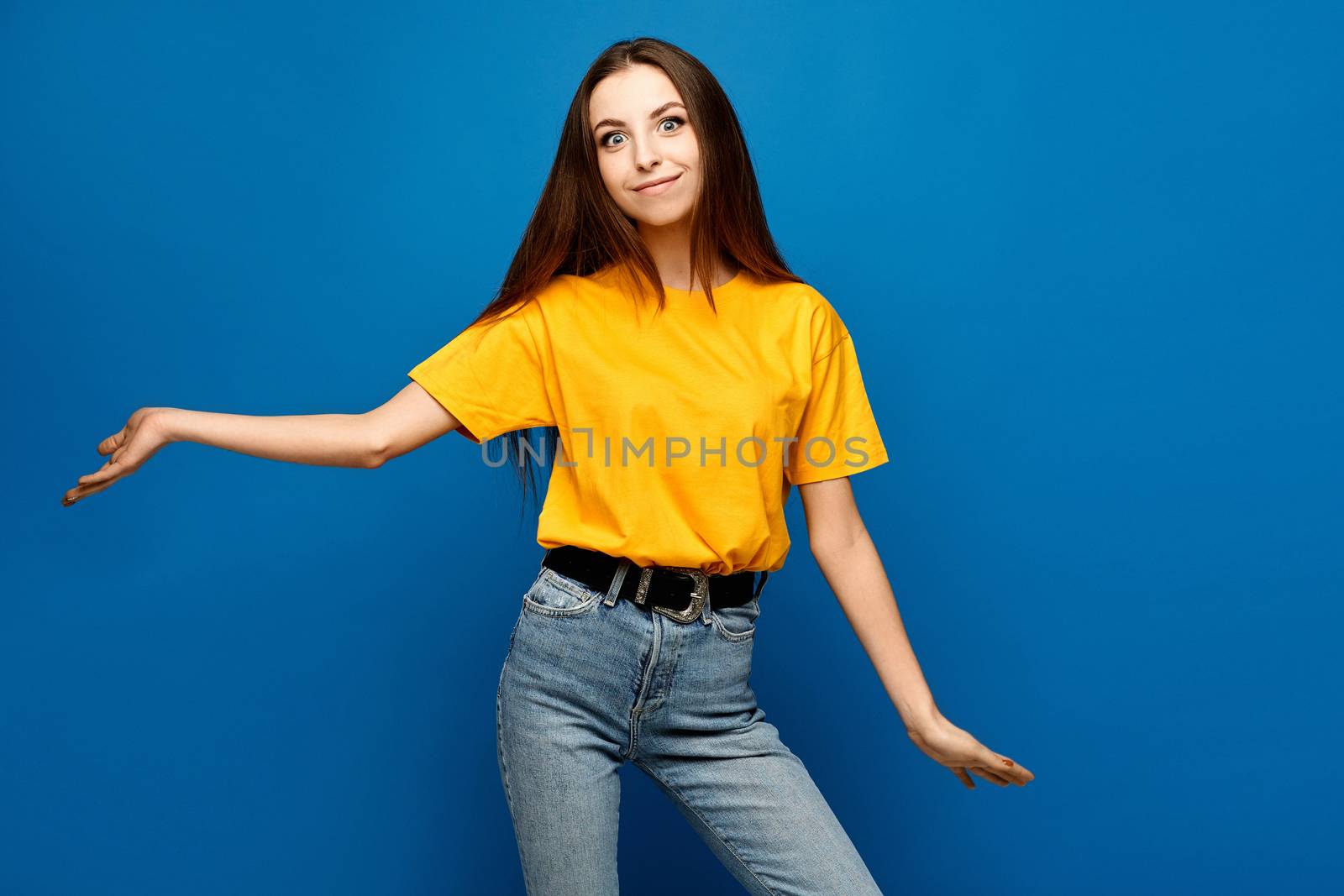 A young surprised woman in yellow blank shirt stands with arms apart over blue background, isolated.