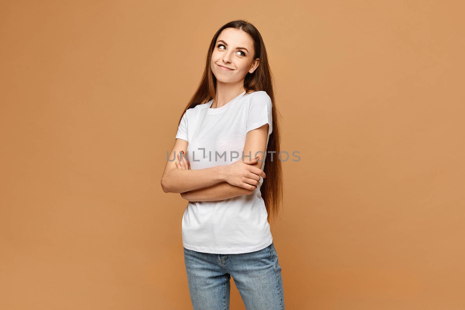 Young woman in white blank t-shirt and blue jeans posing with folded hands at beige background, isolated. Model girl in a casual outfit. Mockup for advertising.