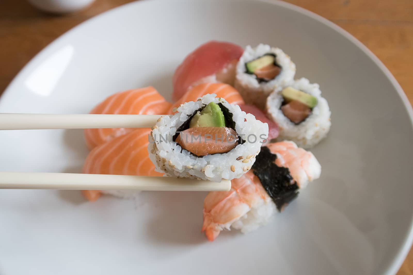 Fresh salmon and avocado uramaki sushi between chopsticks with salmon nigiri, prawn nigiri and tuna nigiri in the background
