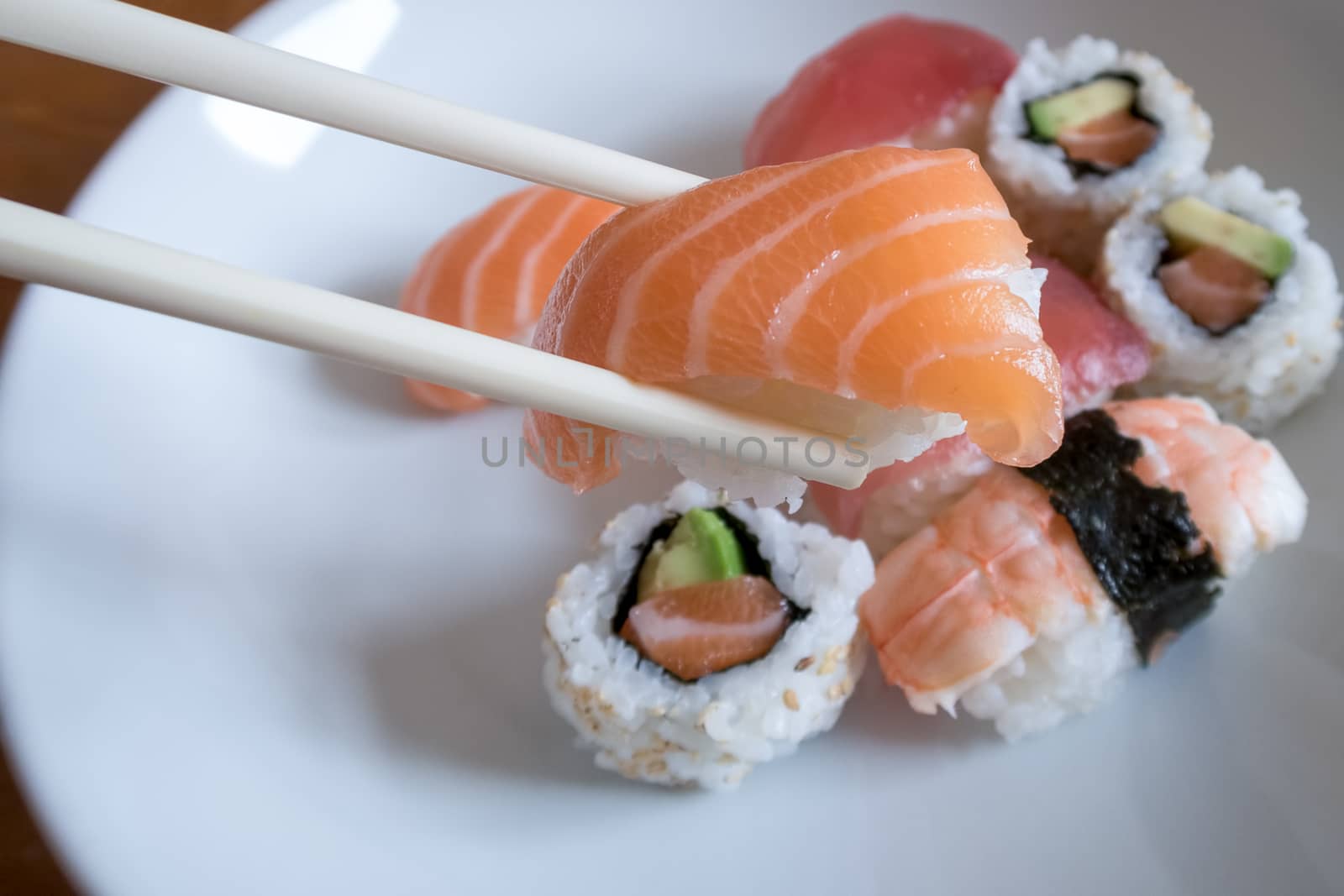 Fresh salmon nigiri sushi between chopsticks with uramaki, prawn nigiri and tuna nigiri in the background