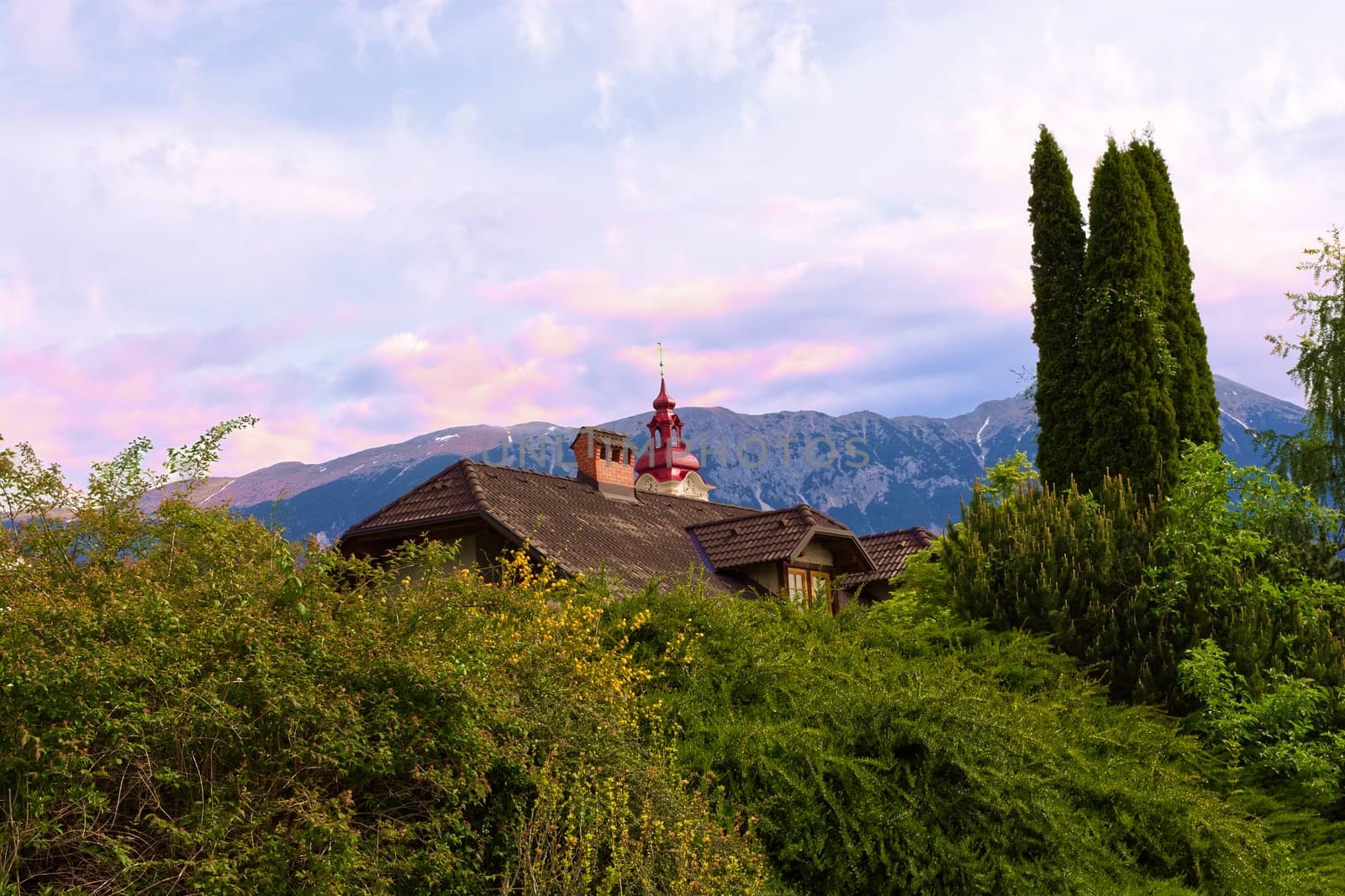 Alpine landscape with a house and a church tower, mountain and sunset on the background