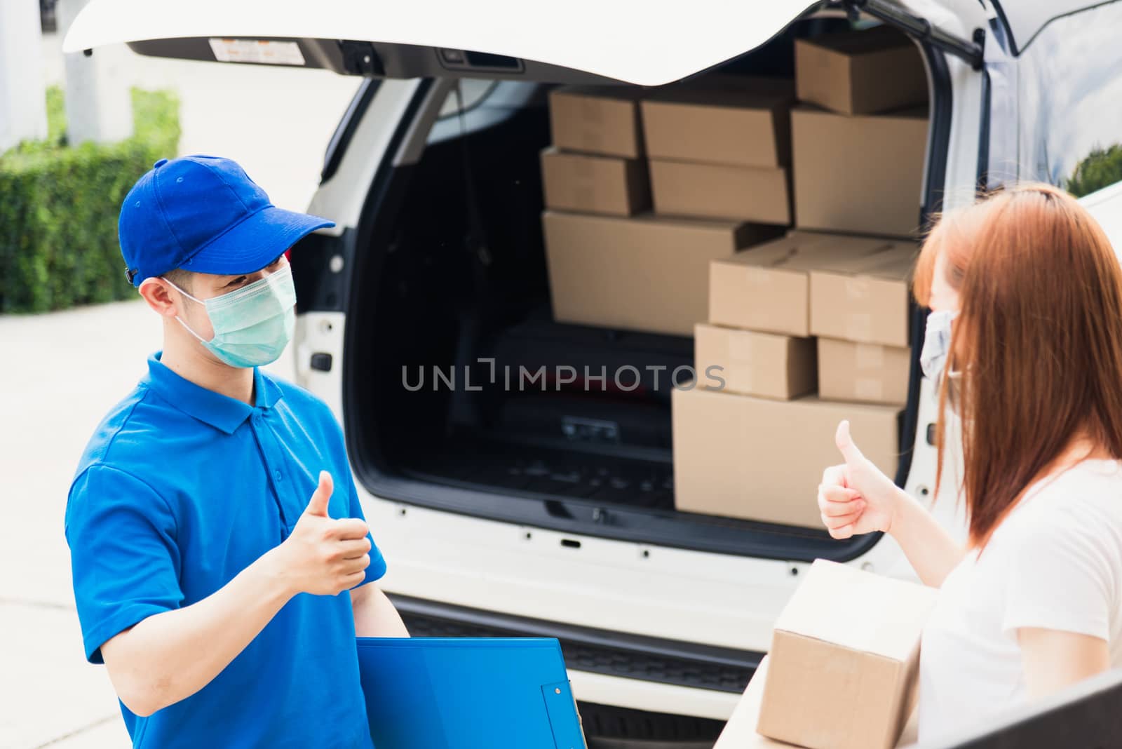 Asian delivery express courier young man giving parcel boxes to woman customer receiving both protective face mask and show thumbs up finger for good support sign, under curfew pandemic coronavirus