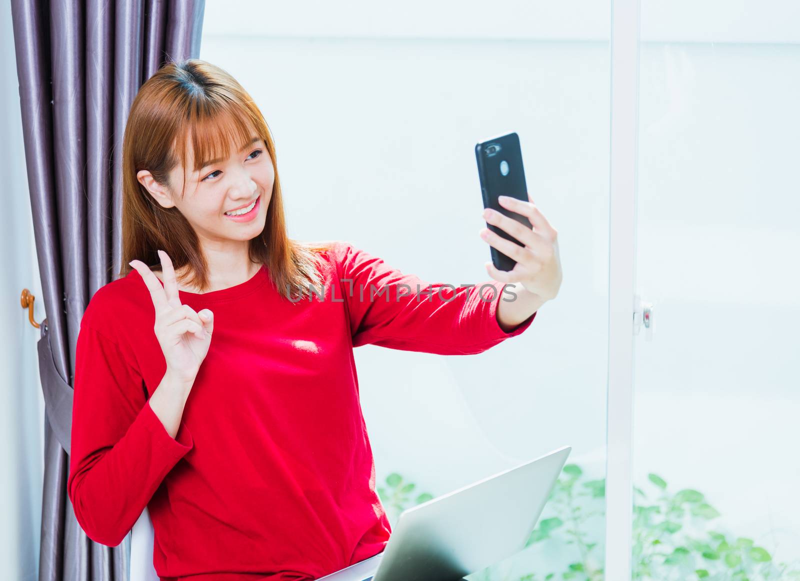 Work from home concept, Asian young woman smiling, student girl sitting on chair make video call or selfie photo from smart mobile phone during working with laptop computer at home office