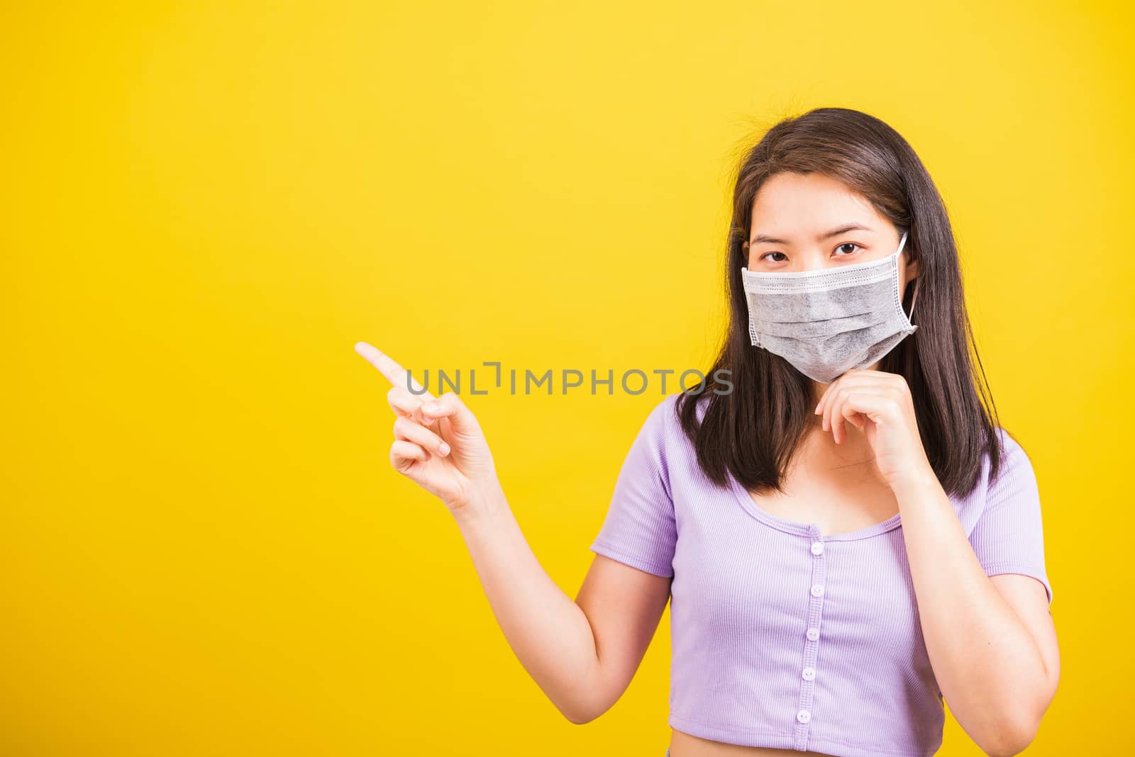 Asian young teen woman wearing face mask protective against coronavirus or COVID-19 virus or filter dust, air pollution her point finger to side space, studio shot isolated yellow background