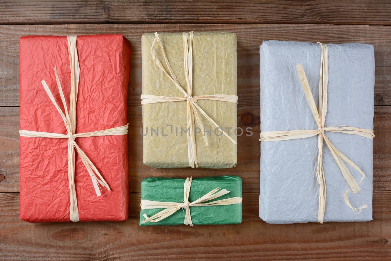 High angle shot of a four presents wrapped with colorful tissue paper. The gifts are tied with raffia and are laying on a rustic wood table. Horizontal format.