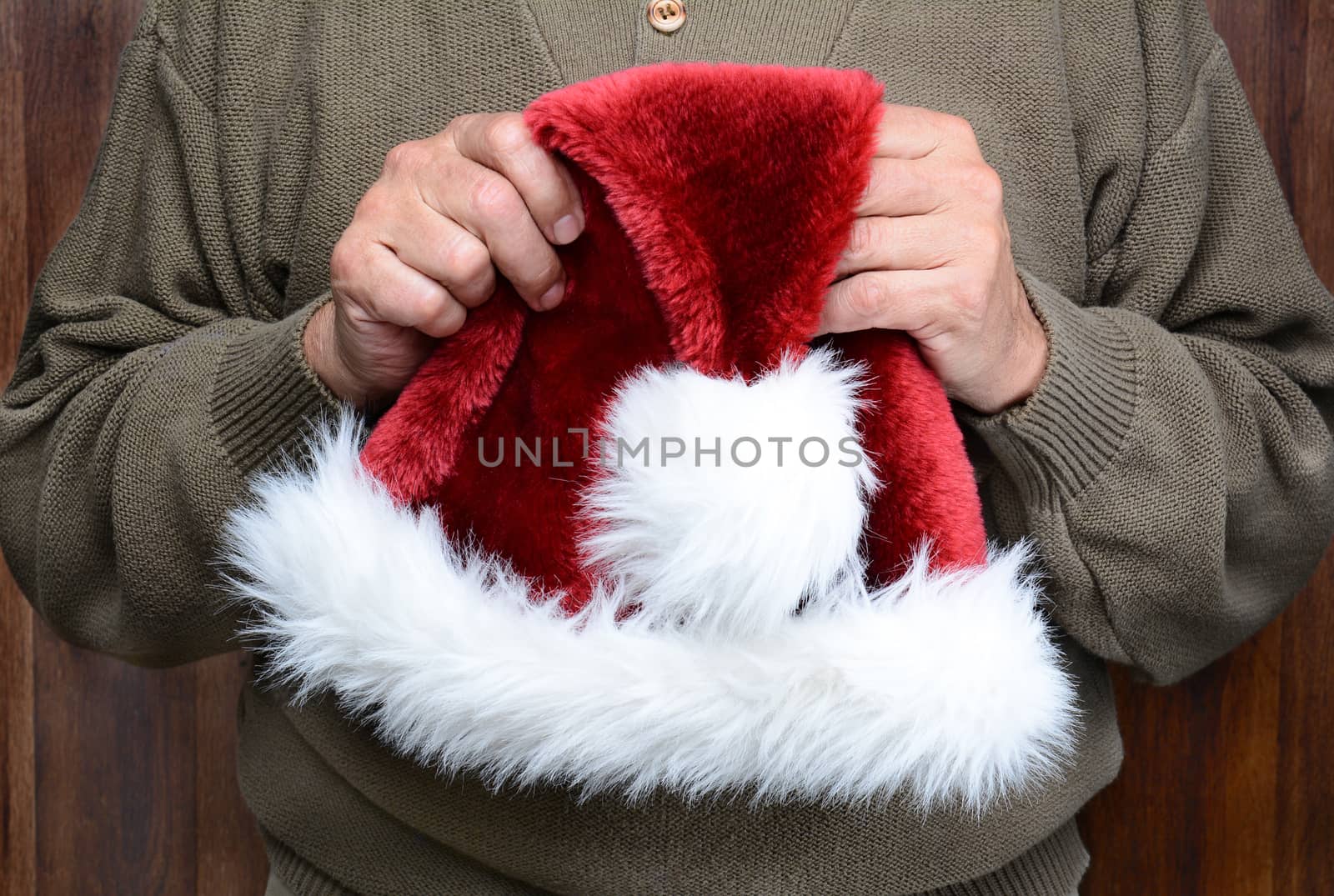 A man wearing a green sweater holding a Santa Claus hat in front of his torso. Man is unrecognizable. Horizontal format.