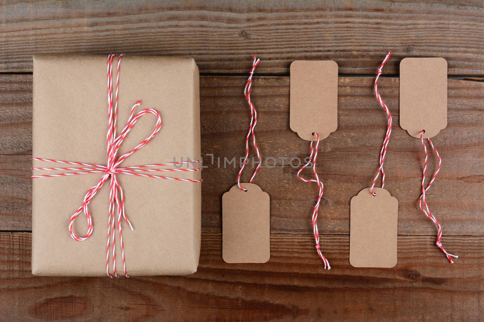 Overhead shot of a Christmas Package wrapped in plain brown paper and tie with red and white string. Four blank tags are next to the gift on a dark rustic wood table.