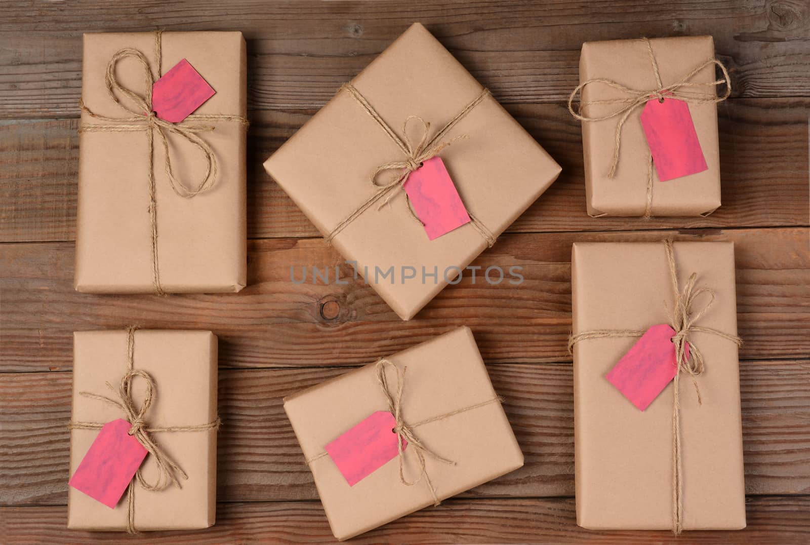 Six holiday packages wrapped in brown kraft paper and tied with twine. The parcels are all different sizes and shapes and resting on a rustic wooden table. There is a blank gift tag attached to each gift. Shot form a high angle.