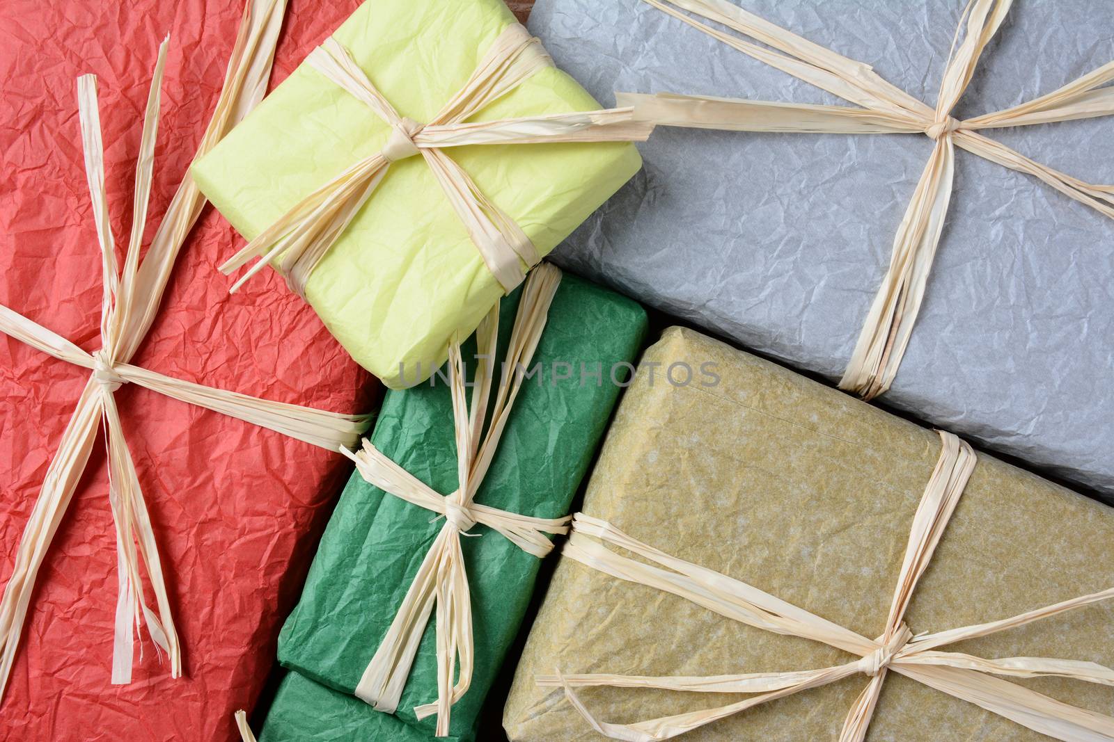 High angle closeup shot of a group of Christmas presents wrapped with colorful tissue paper. The gifts are tied with raffia. Horizontal format filling the frame.