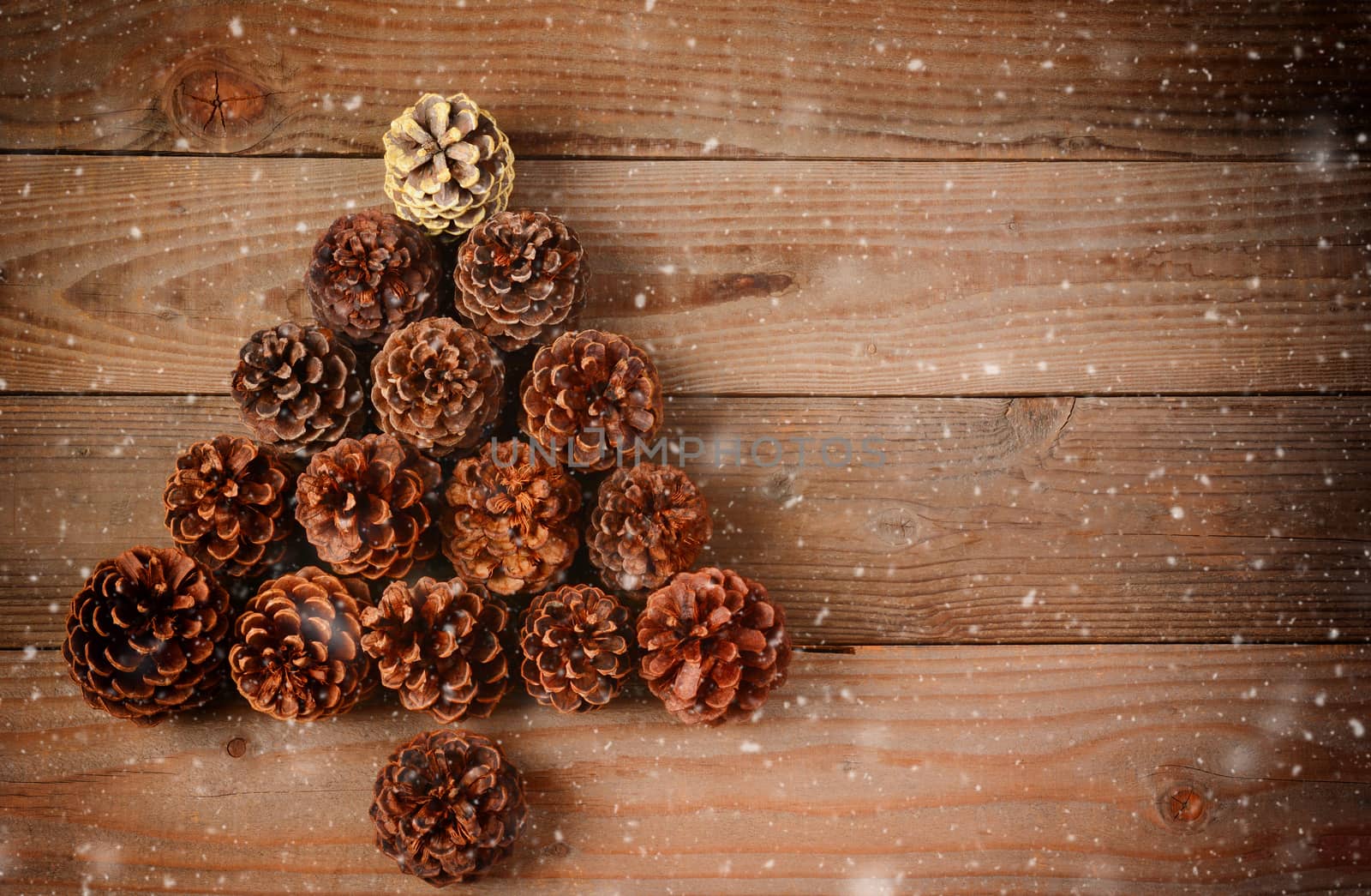 A group of pine cones arranged in a Christmas Tree shape on a rustic wood floor with snow flakes effect. Horizontal format with an instagram retro look and copy space.