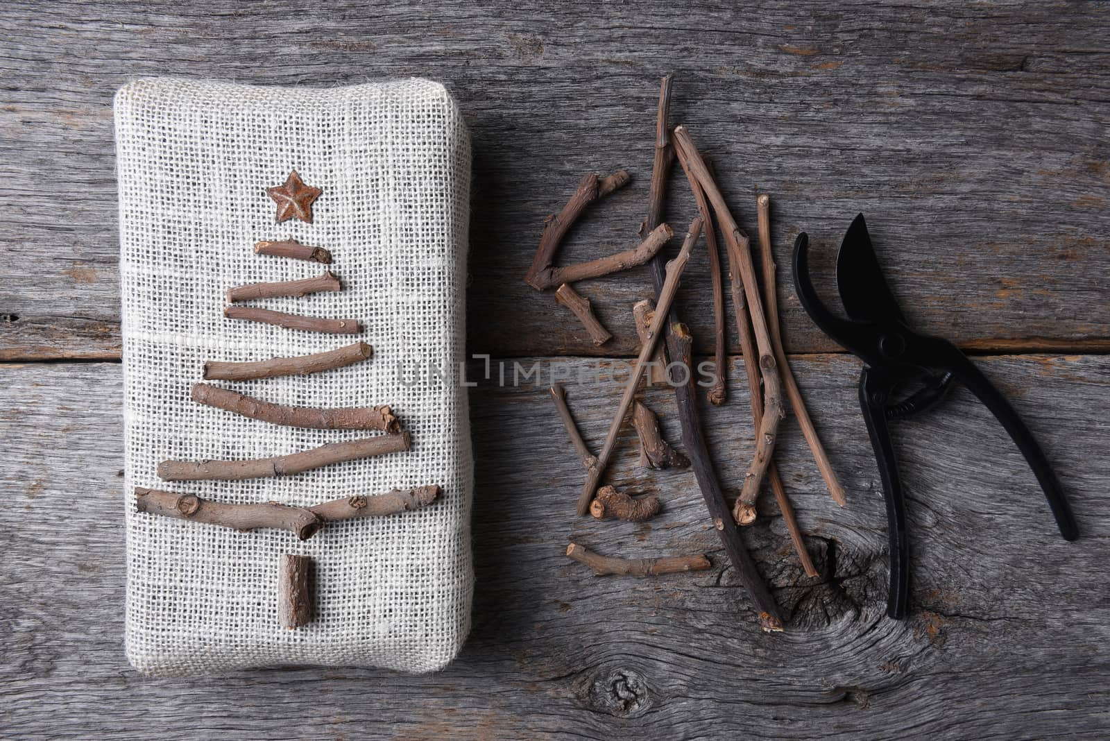 Overhead shot of a burlap wrapped Christmas present with a twig tree and star. Assorted twigs and pruning shears are also on the rustic wood table.