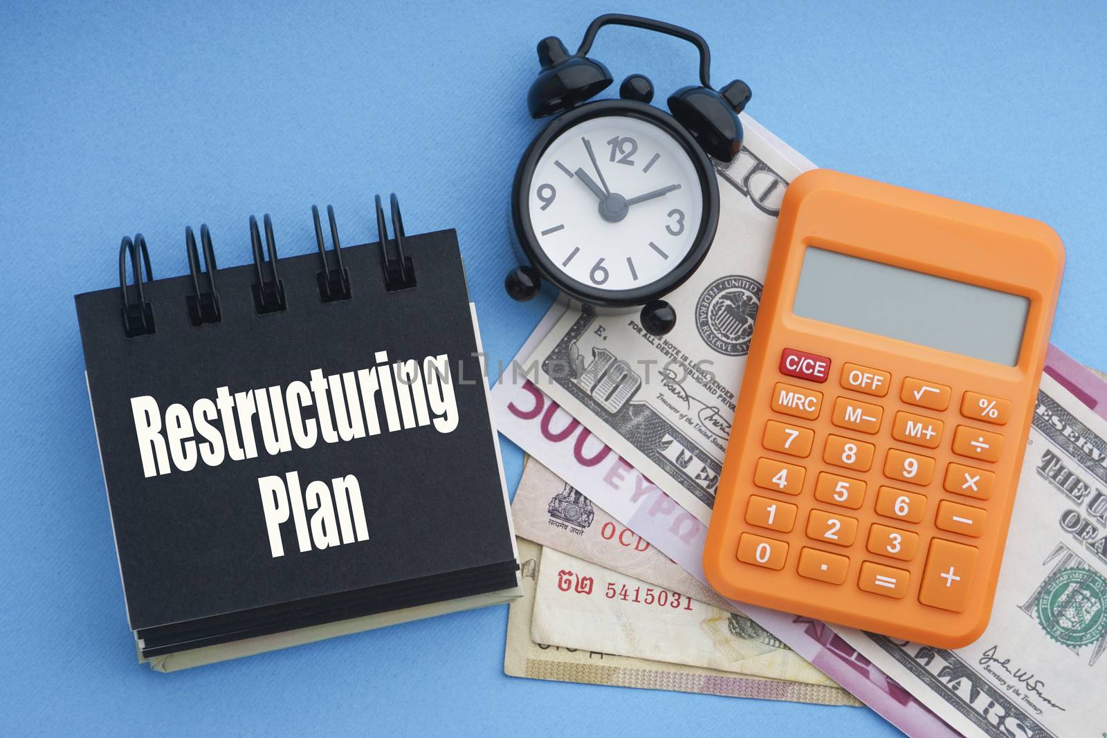 RESTRUCTURING PLAN text with alarm clock, banknotes currencies, notepad and calculator on blue background. Coronavirus Covid19 and Business Concept