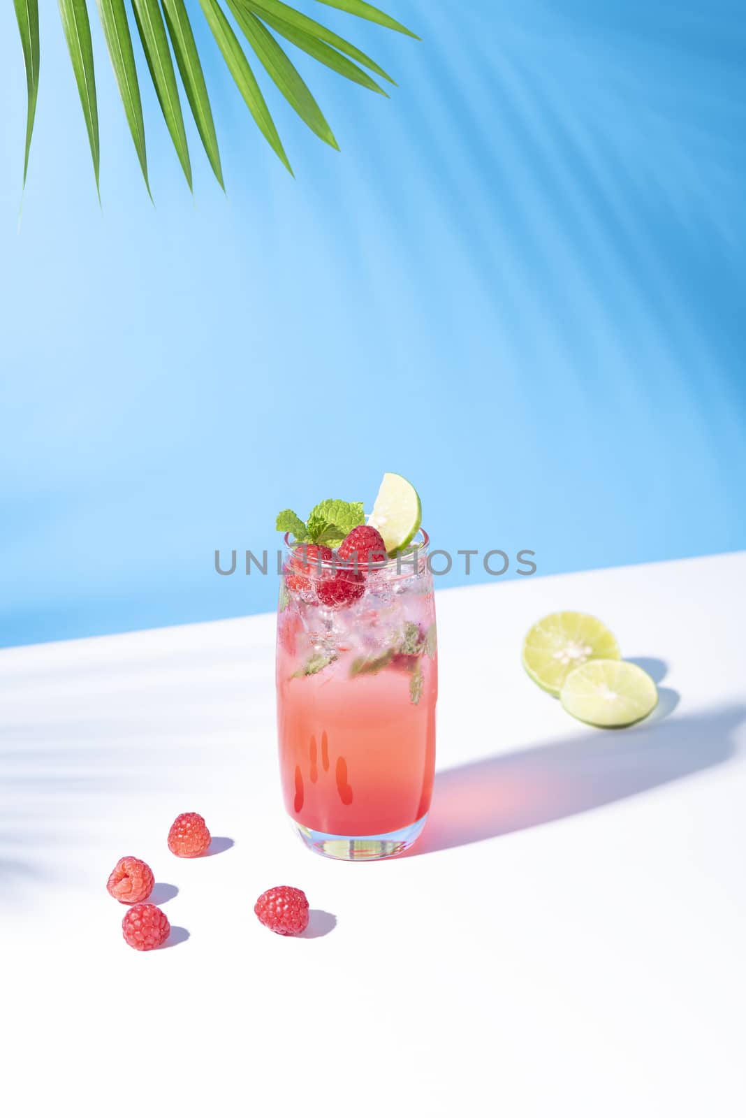 iced red raspberry punch cocktail with lime in glass on blue background. summer drink.