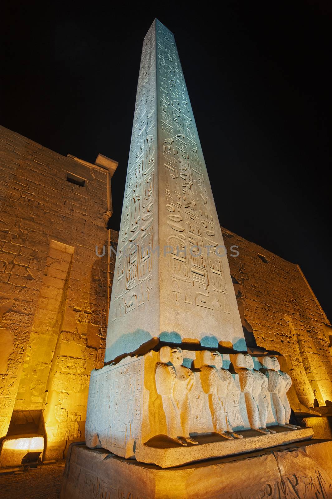 Large obelisk with statues at entrance pylon to ancient egyptian Luxor Temple lit up during night
