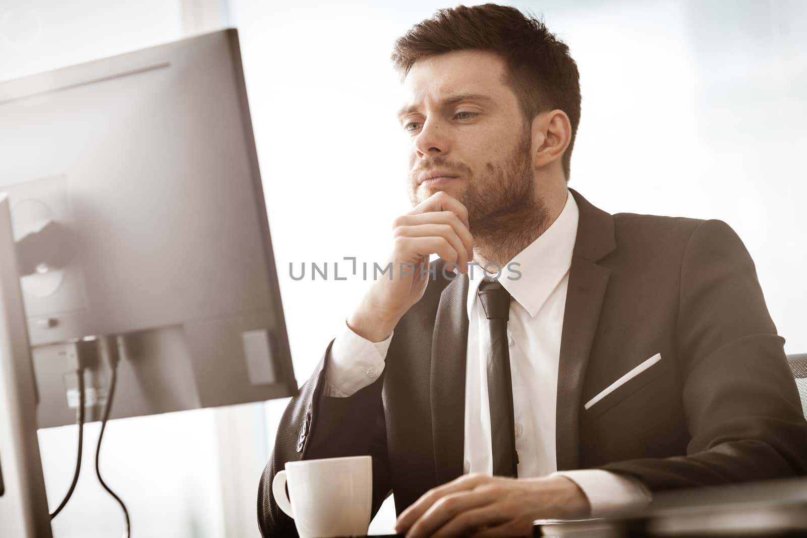 Business crisis concept. Young businessman sitting at the office table busy talking on a cell phone resolving a very serious work problem. Man in suit indoors on glass window background.