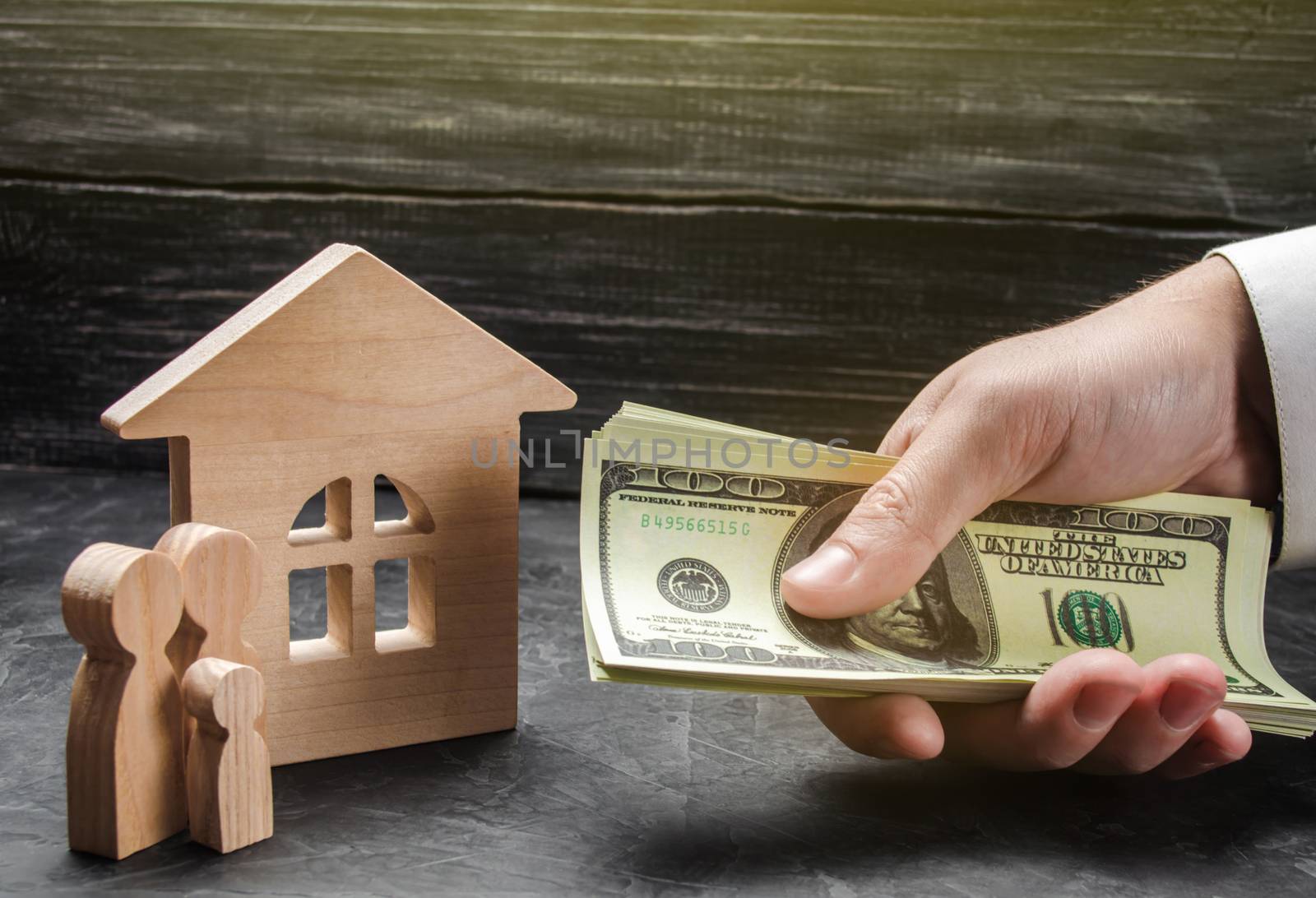 The hand of a businessman stretches a wad of money to the family figures near the wooden house. Providing a mortgage loan for buying a home and improving home conditions. Buying a house