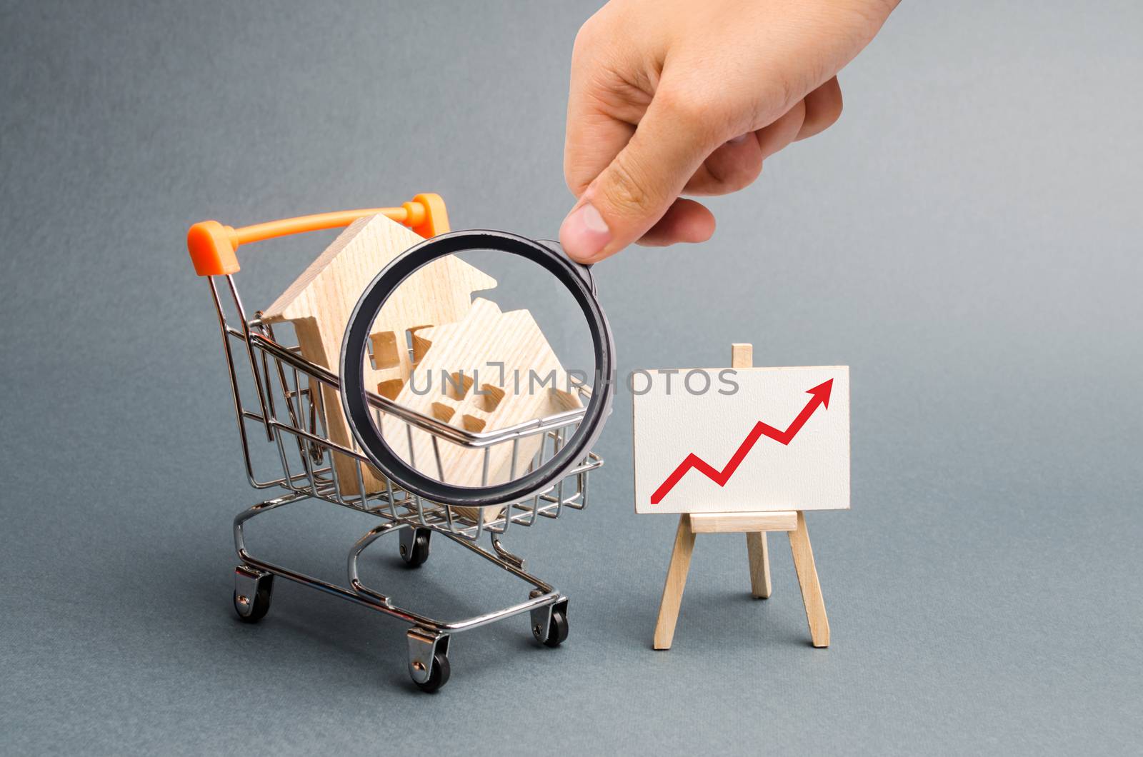 Magnifying glass is looking at the Wooden houses in a supermarket cart and red up arrow. Growing demand for housing and real estate. The growth of population. Investments. Selective focus